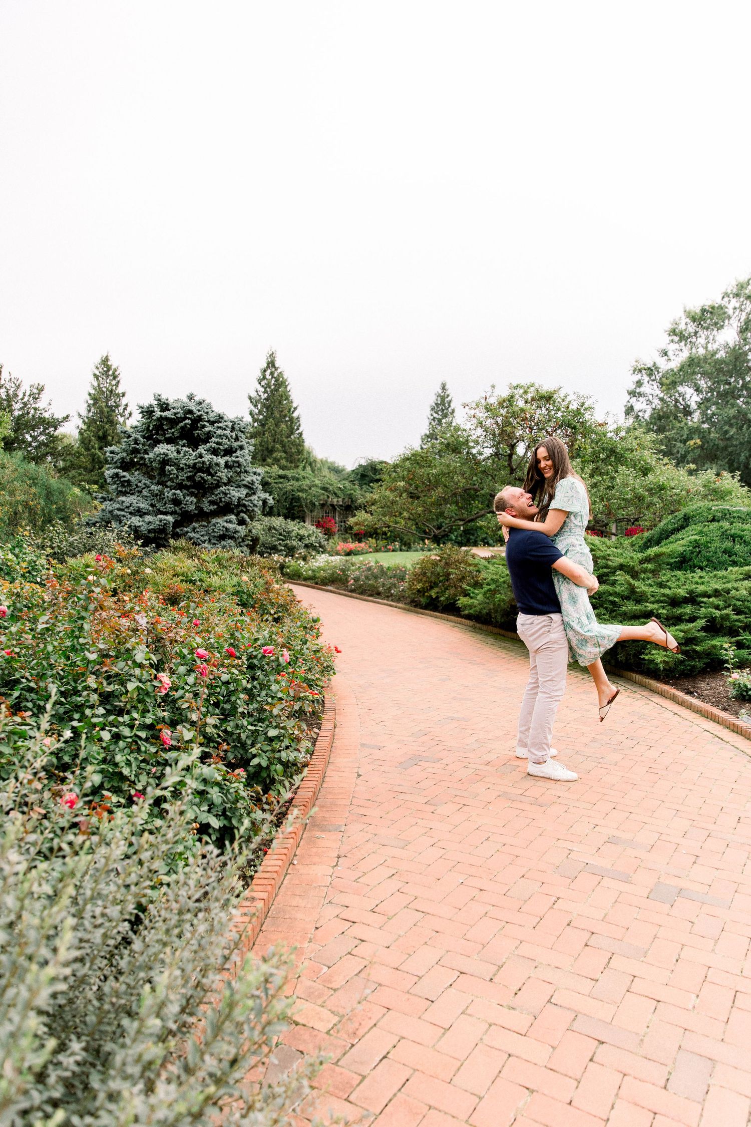 A joyful couple embraces on a brick garden path surrounded by lush greenery and blooming flowers.