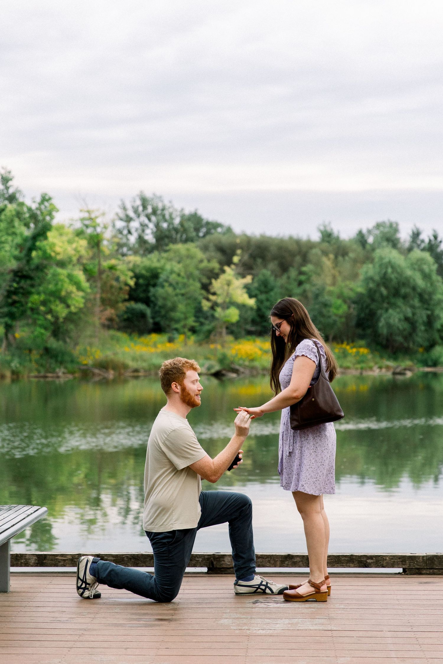 Man kneeling on one knee proposing to woman on wooden dock by scenic lake surrounded by lush green trees.
