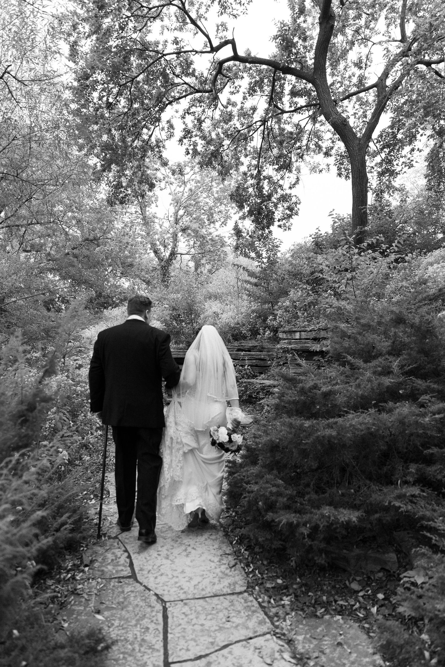 Bride and groom walk hand-in-hand along a lush garden stone path, captured in timeless black and white photography.