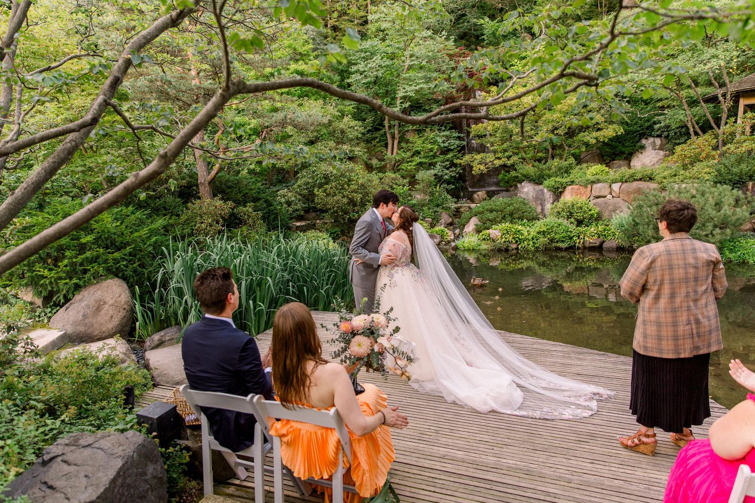 Couple shares first kiss during intimate outdoor wedding ceremony in lush Japanese garden with guests seated on wooden deck.