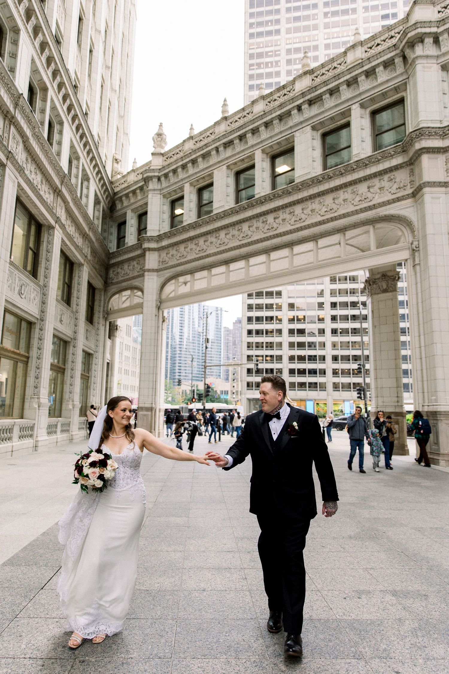 Bride and groom walk hand in hand beneath ornate arch of Wrigley Building in Chicago.