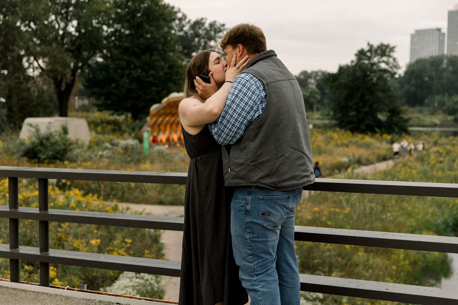 Couple sharing a romantic kiss on a bridge surrounded by lush gardens and city skyline in the background.