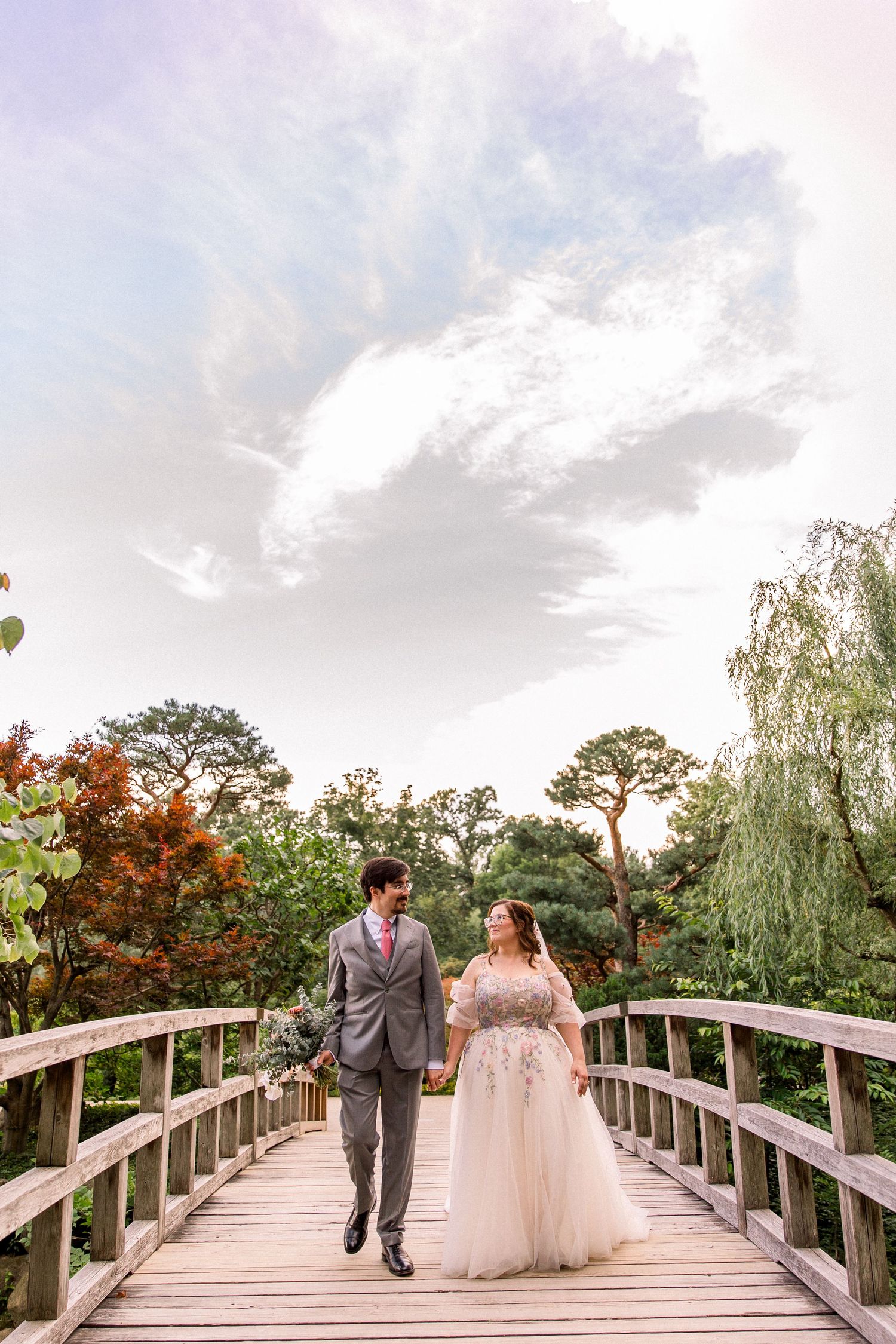 Bride and groom walk hand-in-hand on a wooden bridge surrounded by lush gardens under a dramatic cloudy sky.