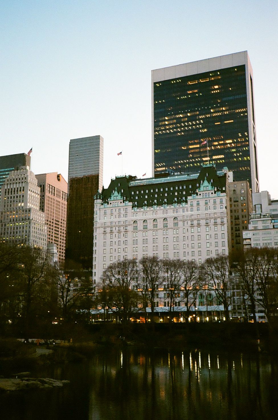 Manhattan skyline at dusk featuring the Plaza Hotel and modern skyscrapers reflected in Central Park pond.
