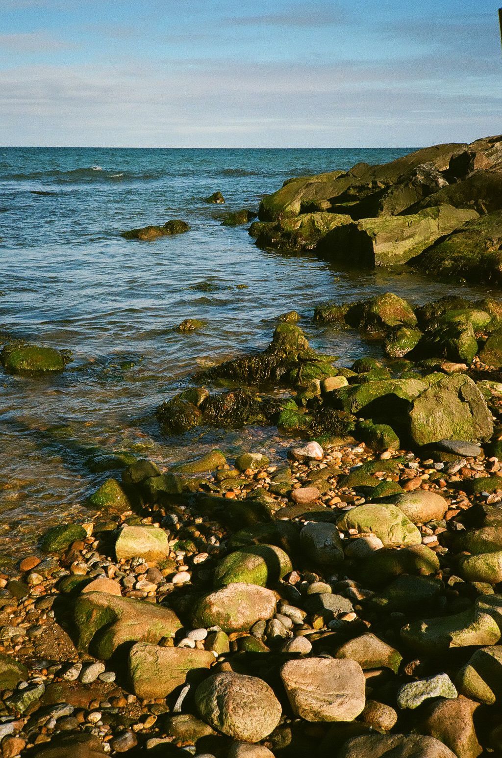 Rocky coastline with waves crashing against moss-covered boulders under cloudy skies.
