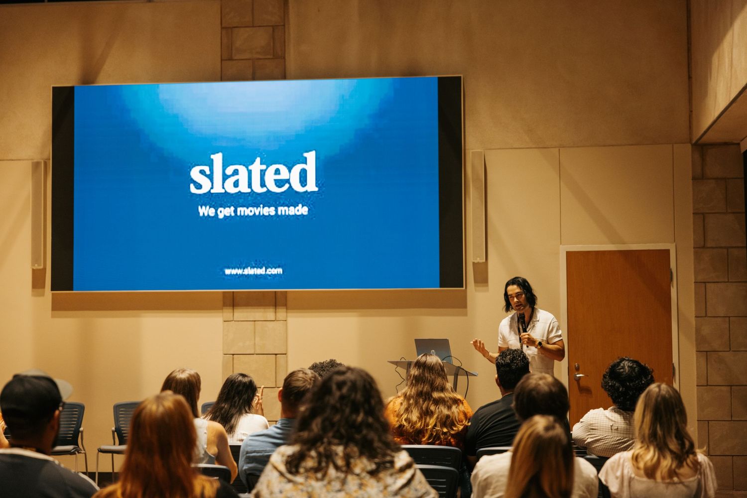 A presenter stands at the front of a classroom with a blue screen displaying the Slated logo behind them.