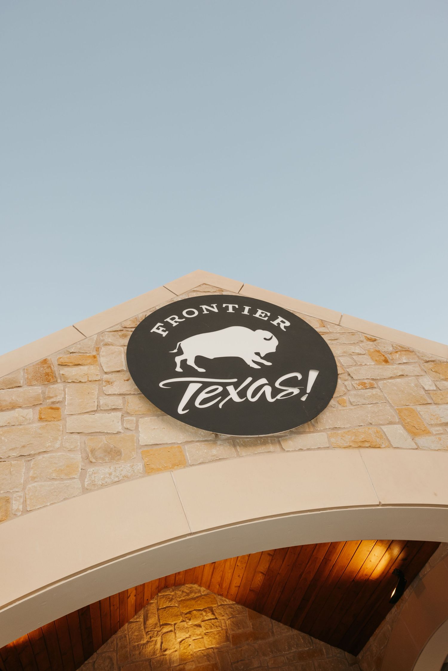 A curved stone wall entrance with a black circular sign featuring the Frontier Texas logo and a buffalo silhouette.
