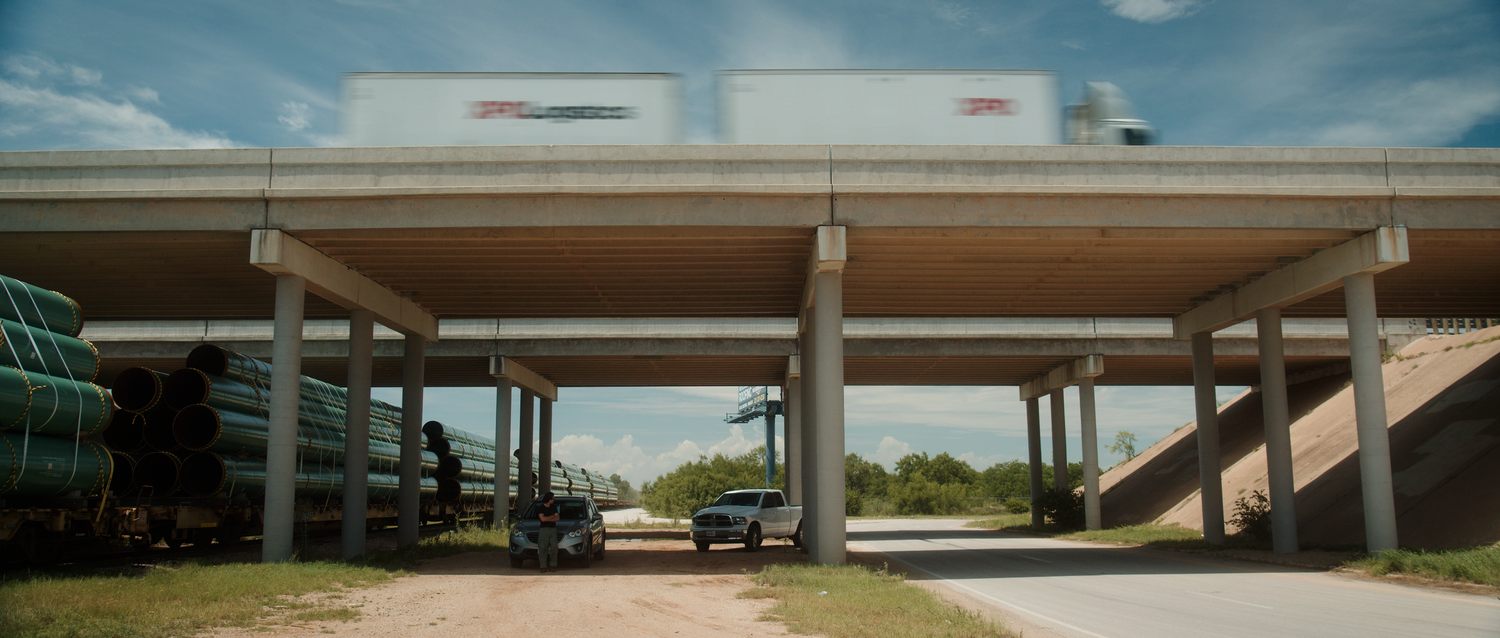 Trucks passing under a concrete highway overpass with green pipes and vehicles on a sunny day.