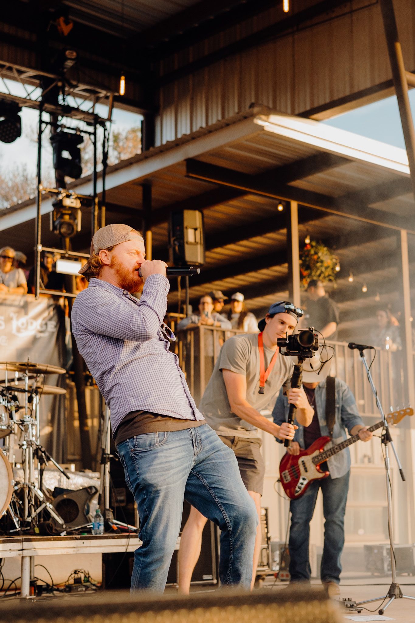 Live band performance on outdoor stage with vocalist singing into mic and guitarist playing red bass guitar.