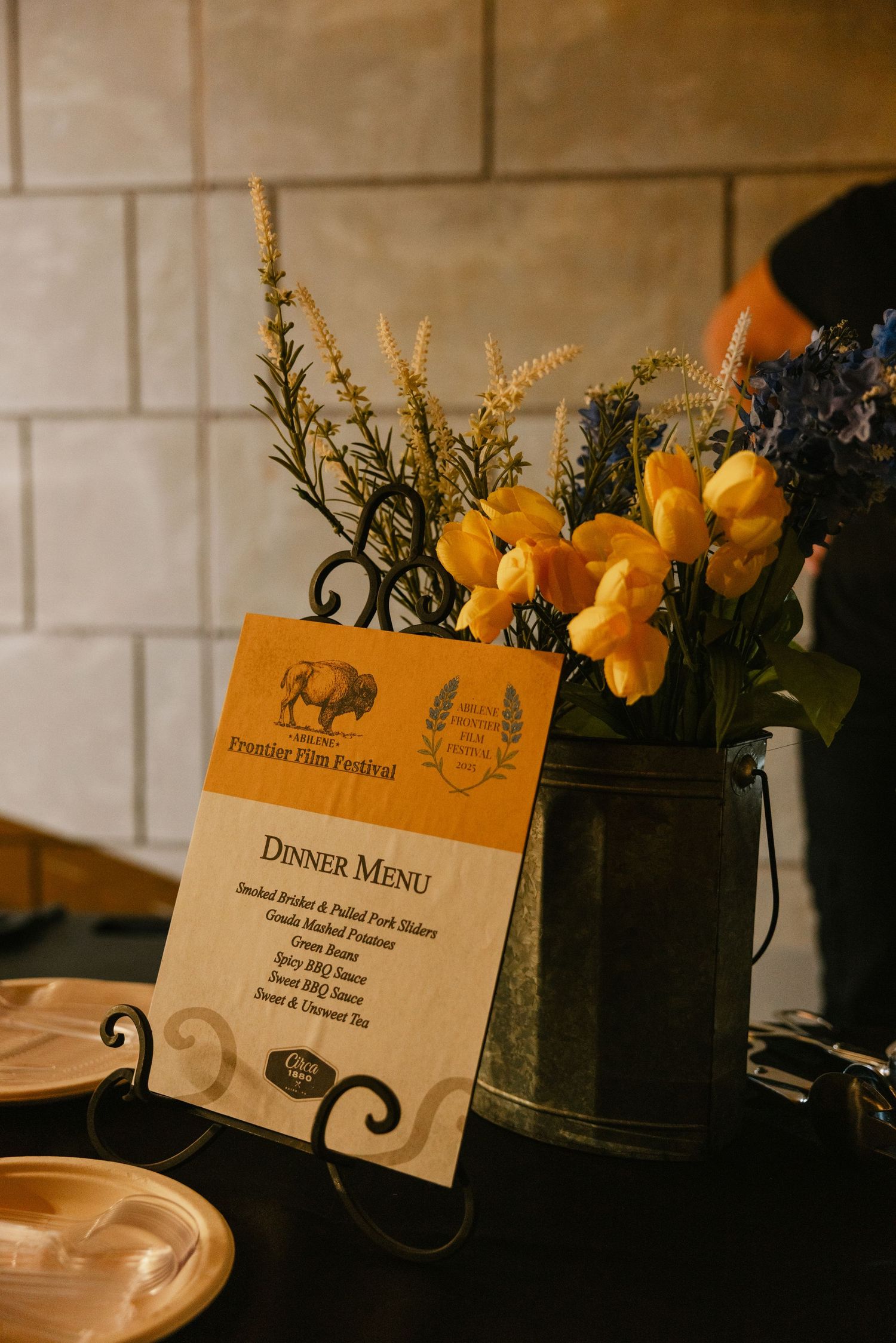 Elegant dinner menu card displayed on a table with yellow flowers and greenery in a dark vase against white brick wall.