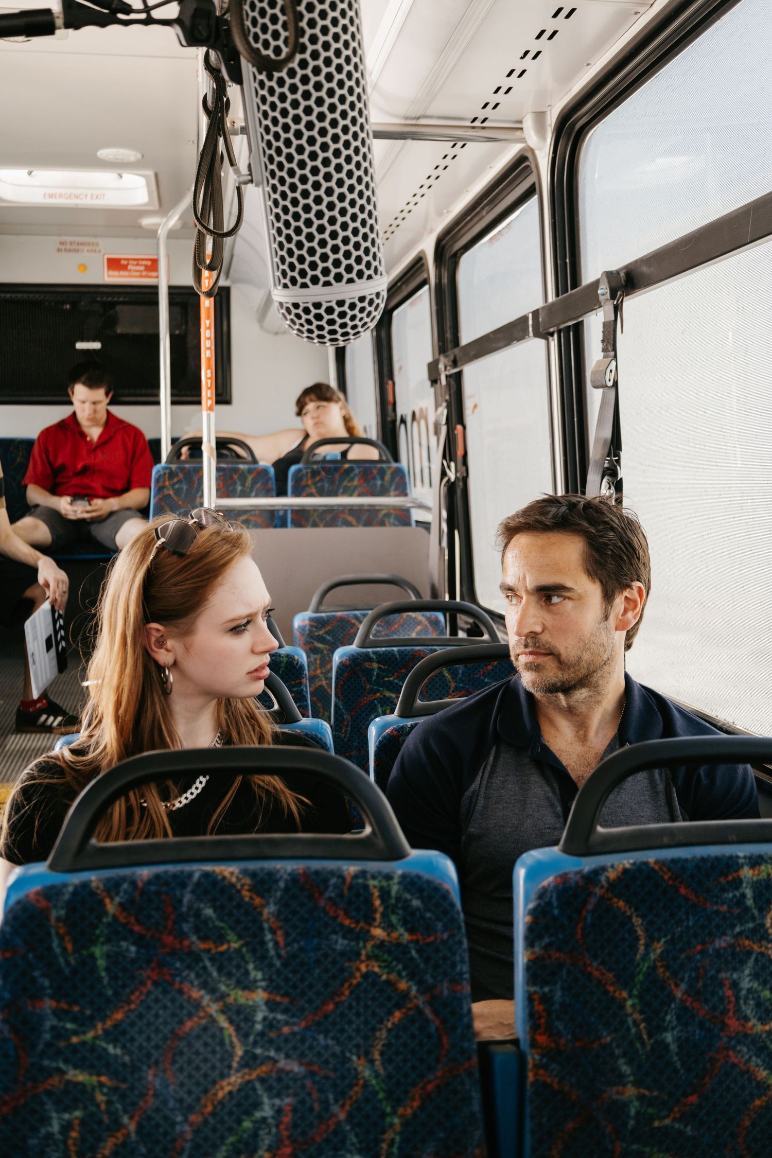 A man and woman sit across from each other on a public bus, engaged in conversation, with other passengers seated behind them.