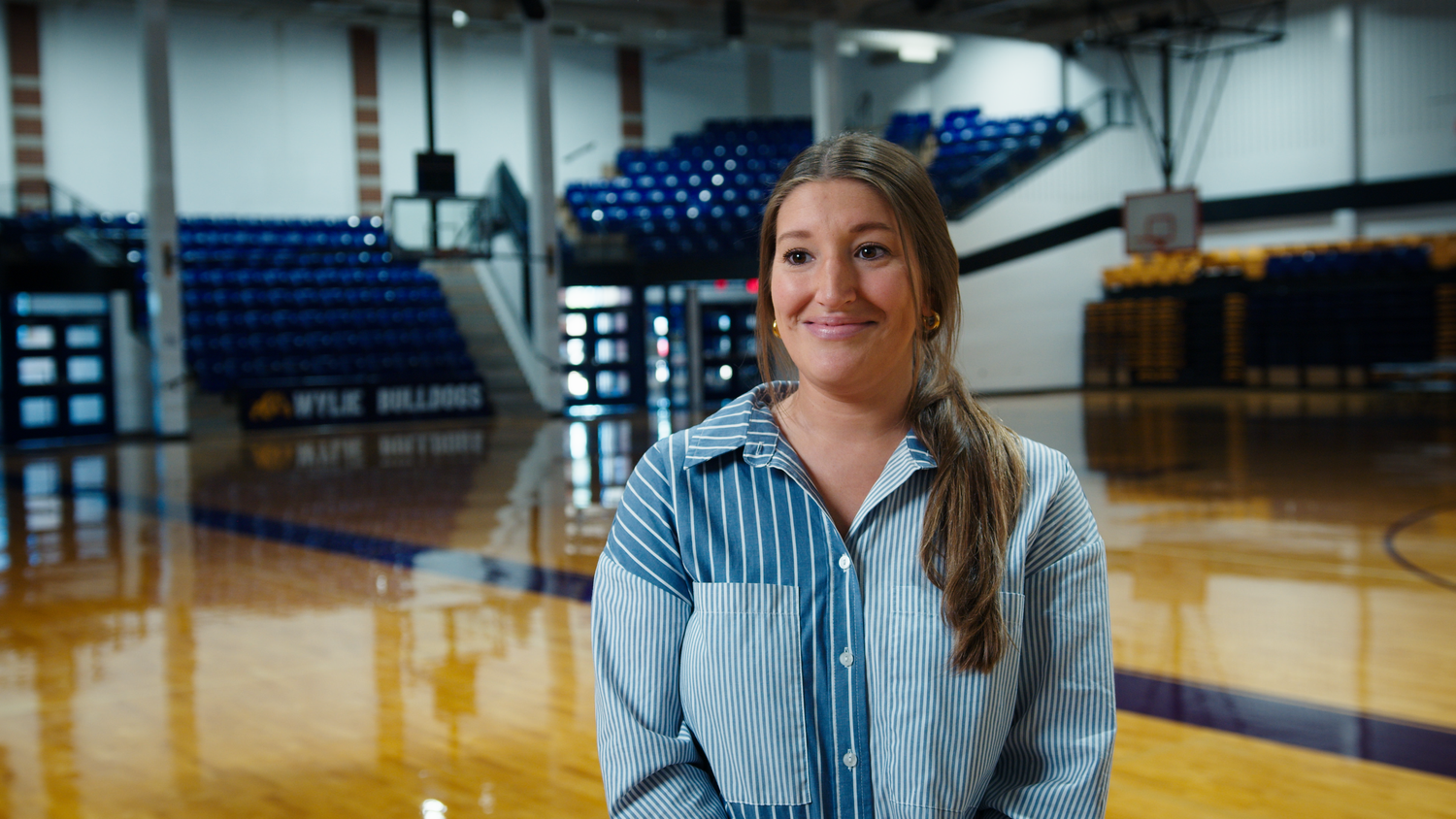 Smiling young woman in a blue striped shirt standing on a basketball court in a school gymnasium.