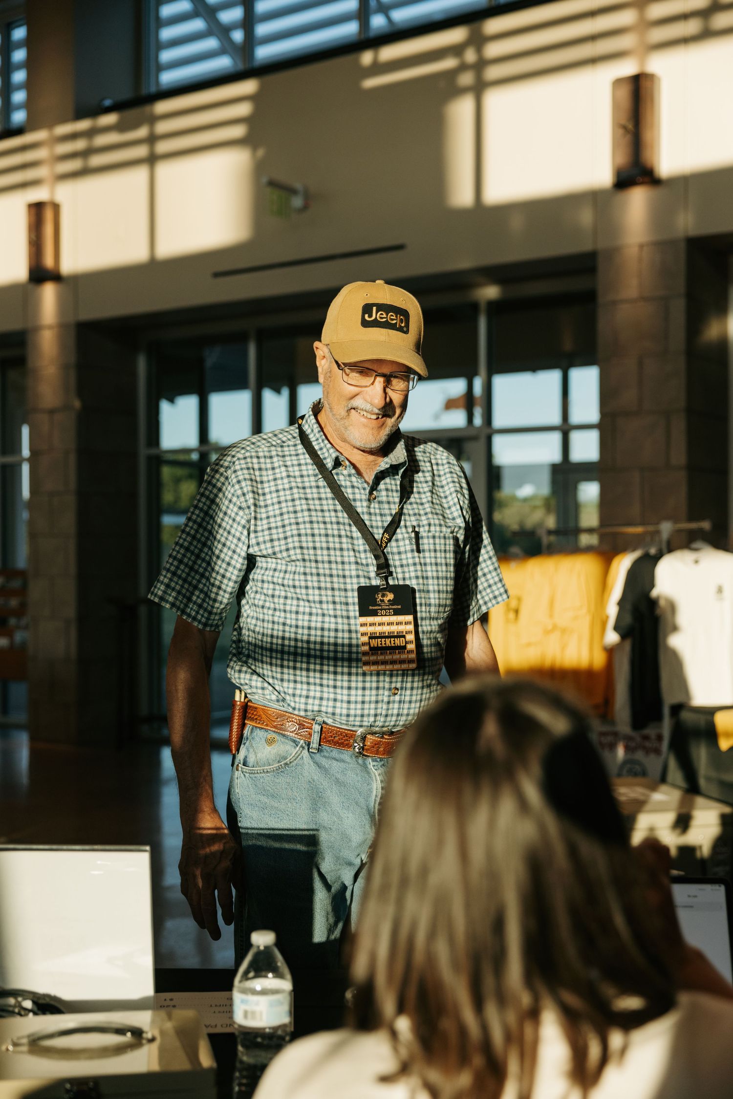 A person in a plaid shirt and baseball cap speaks to an audience in a well-lit indoor venue.