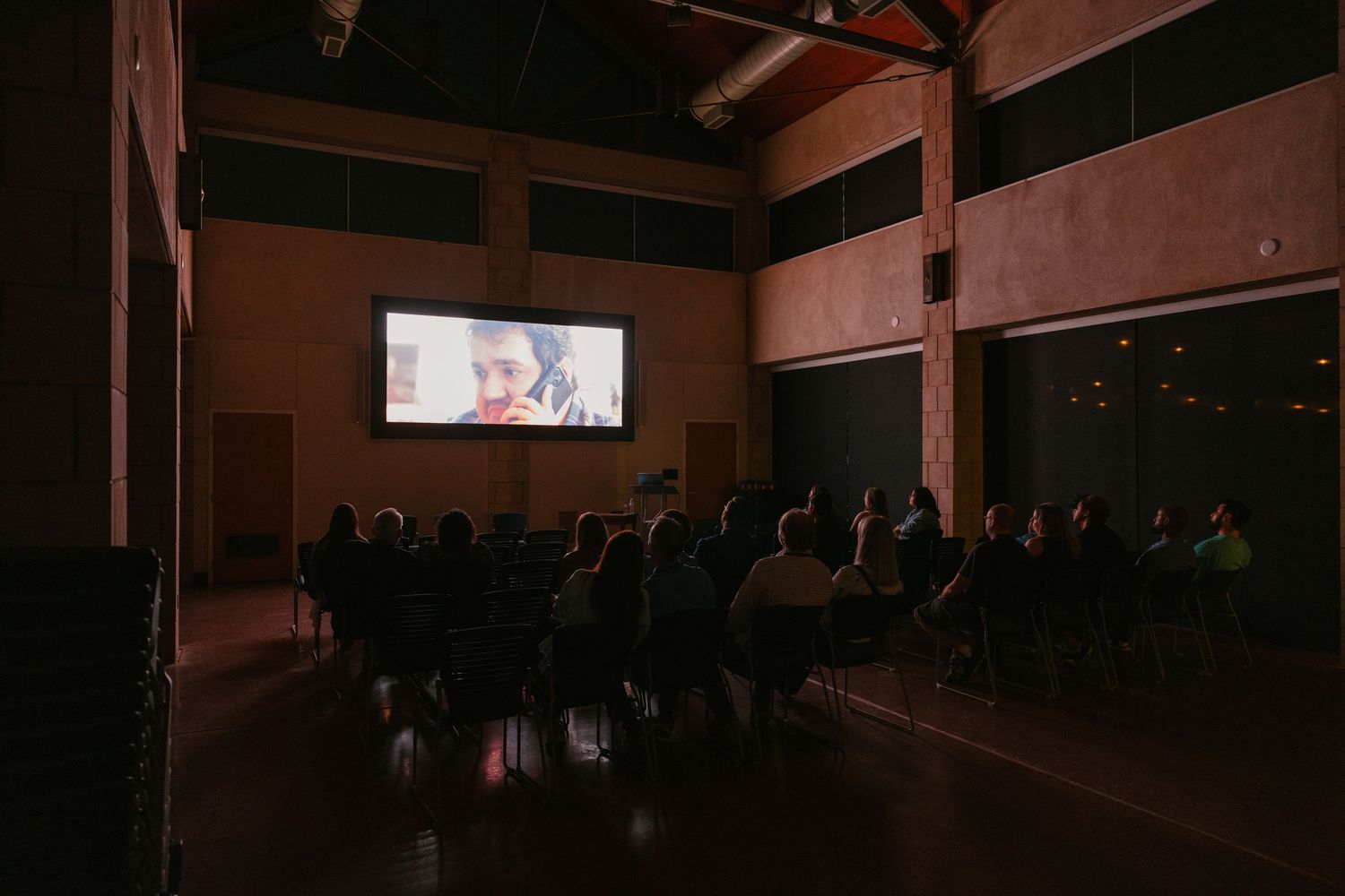 Dark silhouettes of people seated in a theater auditorium with dim lighting and red tones.