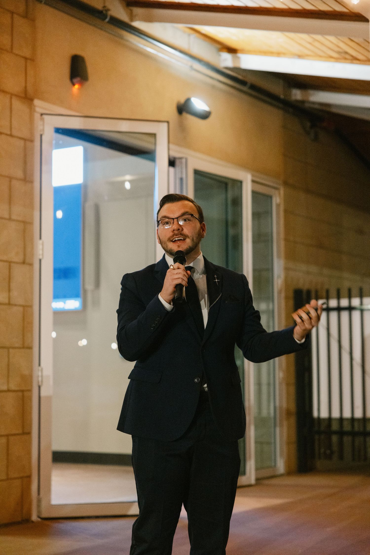 Person in a black suit gestures expressively while speaking in a hallway with wooden ceiling and white walls.