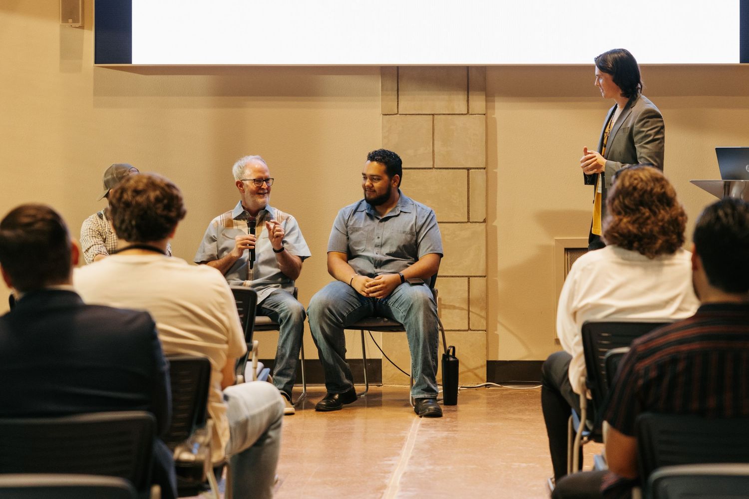 People sit in rows of chairs during an indoor meeting or presentation in a beige-colored room.