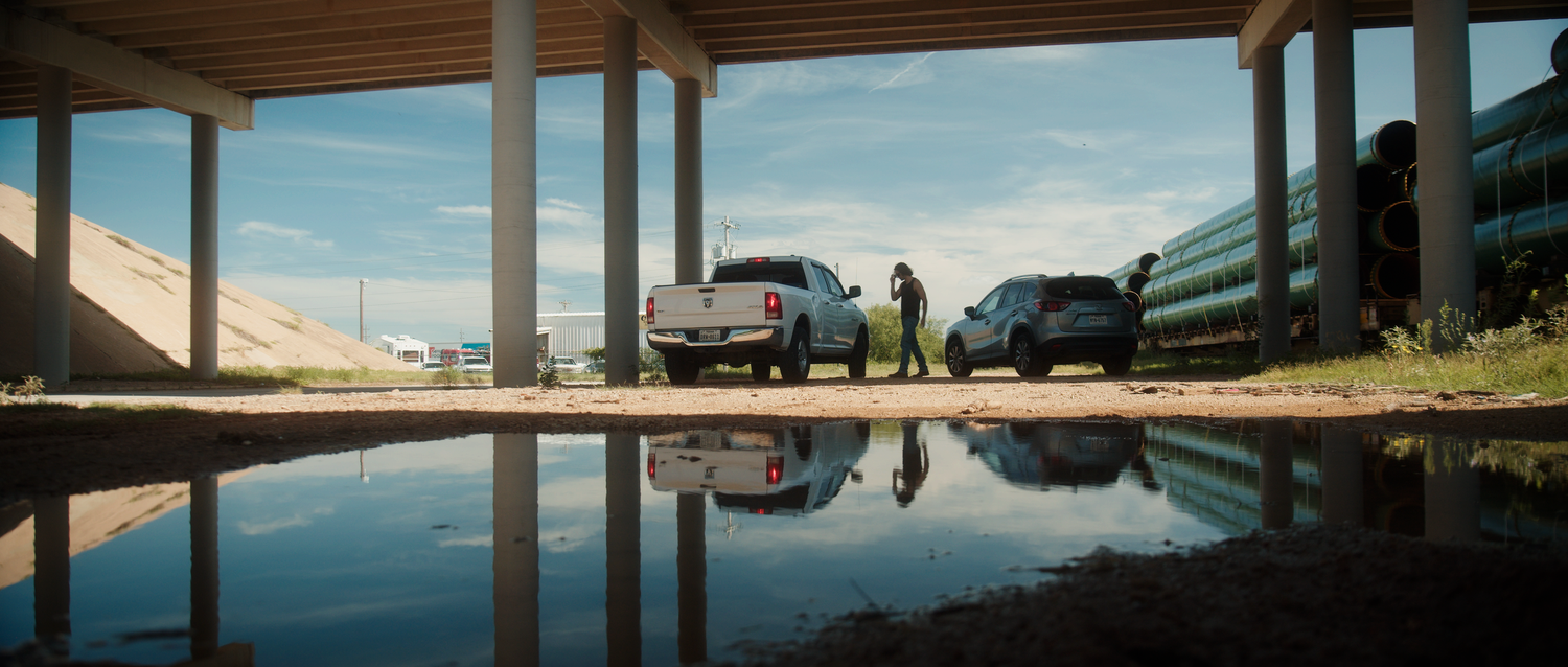 Two vehicles parked under a covered area reflected in a puddle, with a person standing nearby on a sunny day.