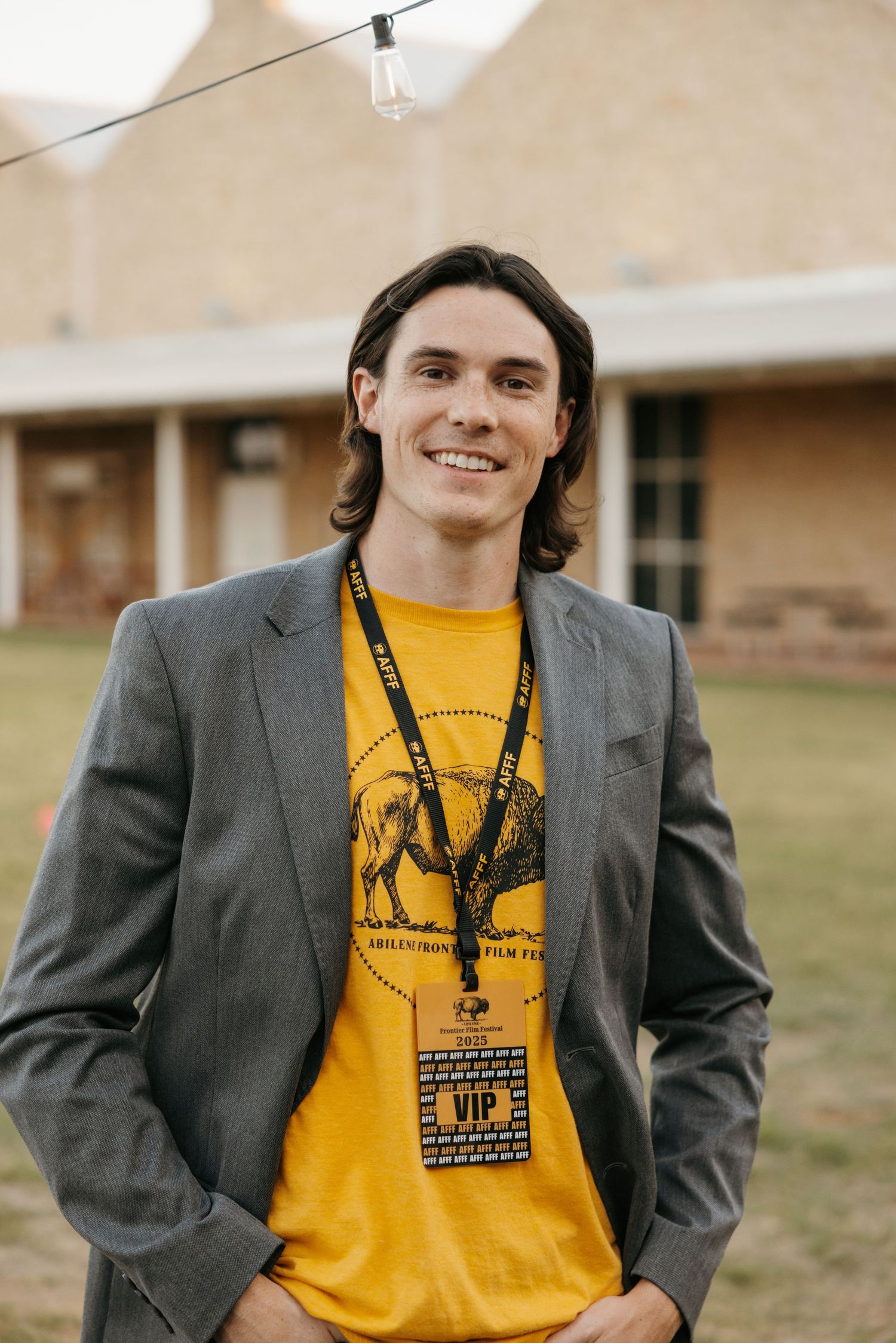 A person in a grey blazer and yellow t-shirt stands outdoors against a brick building background.