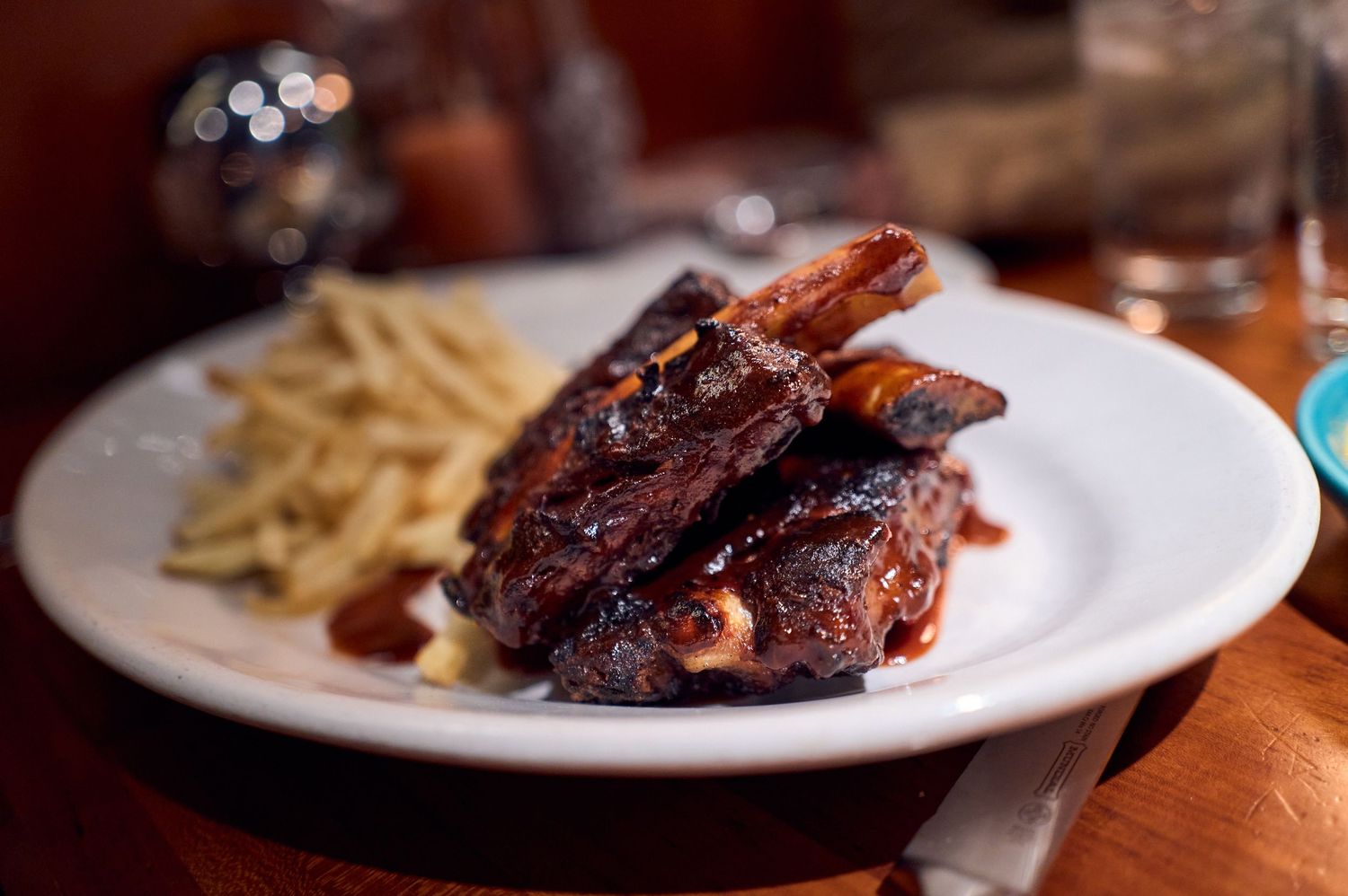 Glazed barbecue ribs with french fries served on a white plate in a restaurant setting.