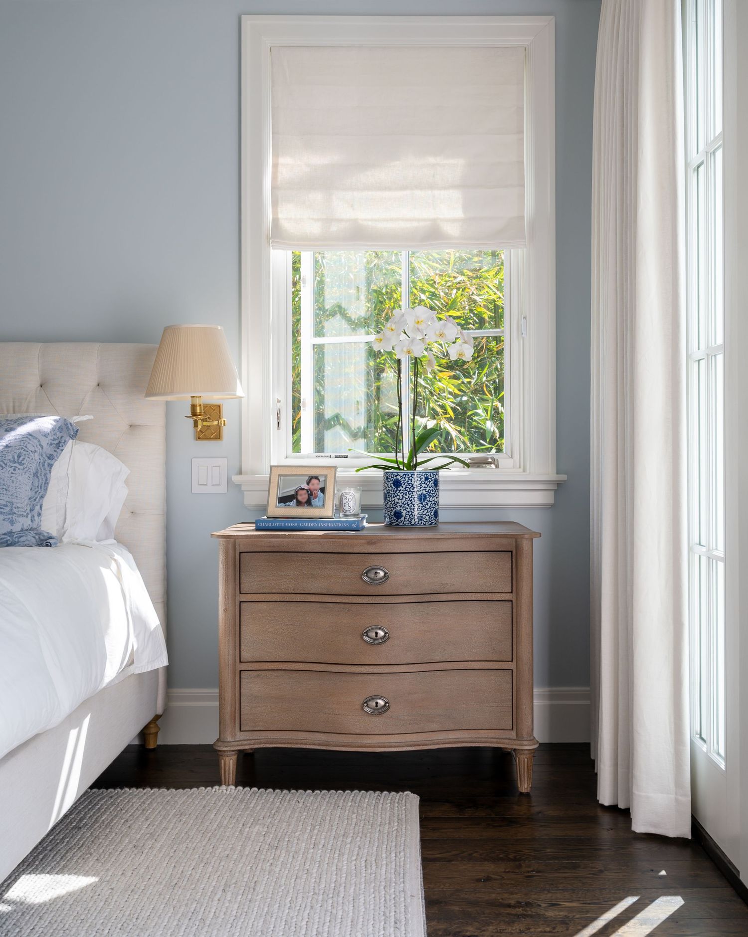 Cozy bedroom corner with white roman shade, wooden nightstand, and natural light streaming through window.