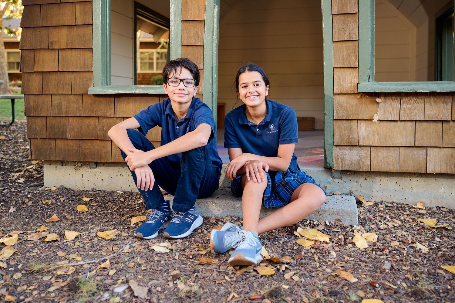 Two students in navy blue uniforms sitting casually on porch steps of a brown shingled building.