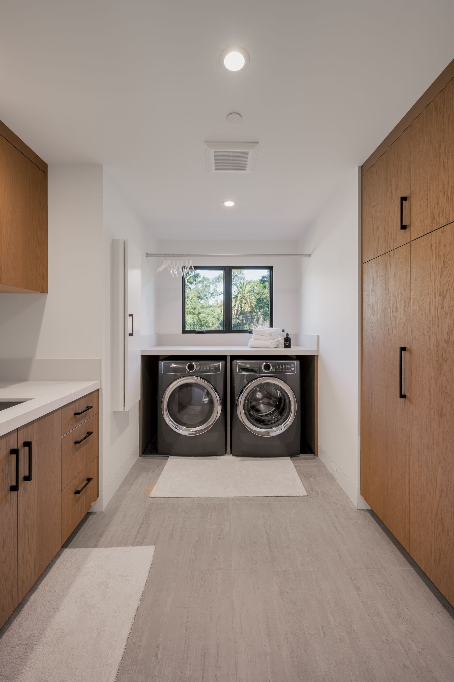 Modern laundry room with wood cabinets, side-by-side washer and dryer, and window overlooking greenery.