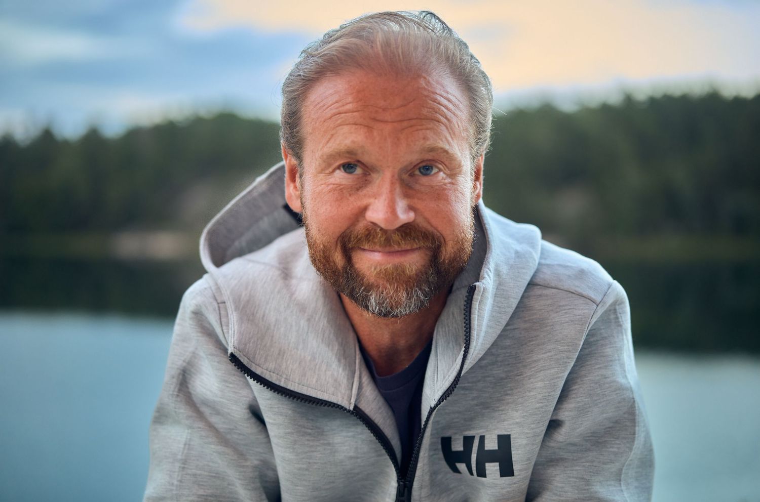 Man in gray Helly Hansen jacket smiles by a lake during sunset.