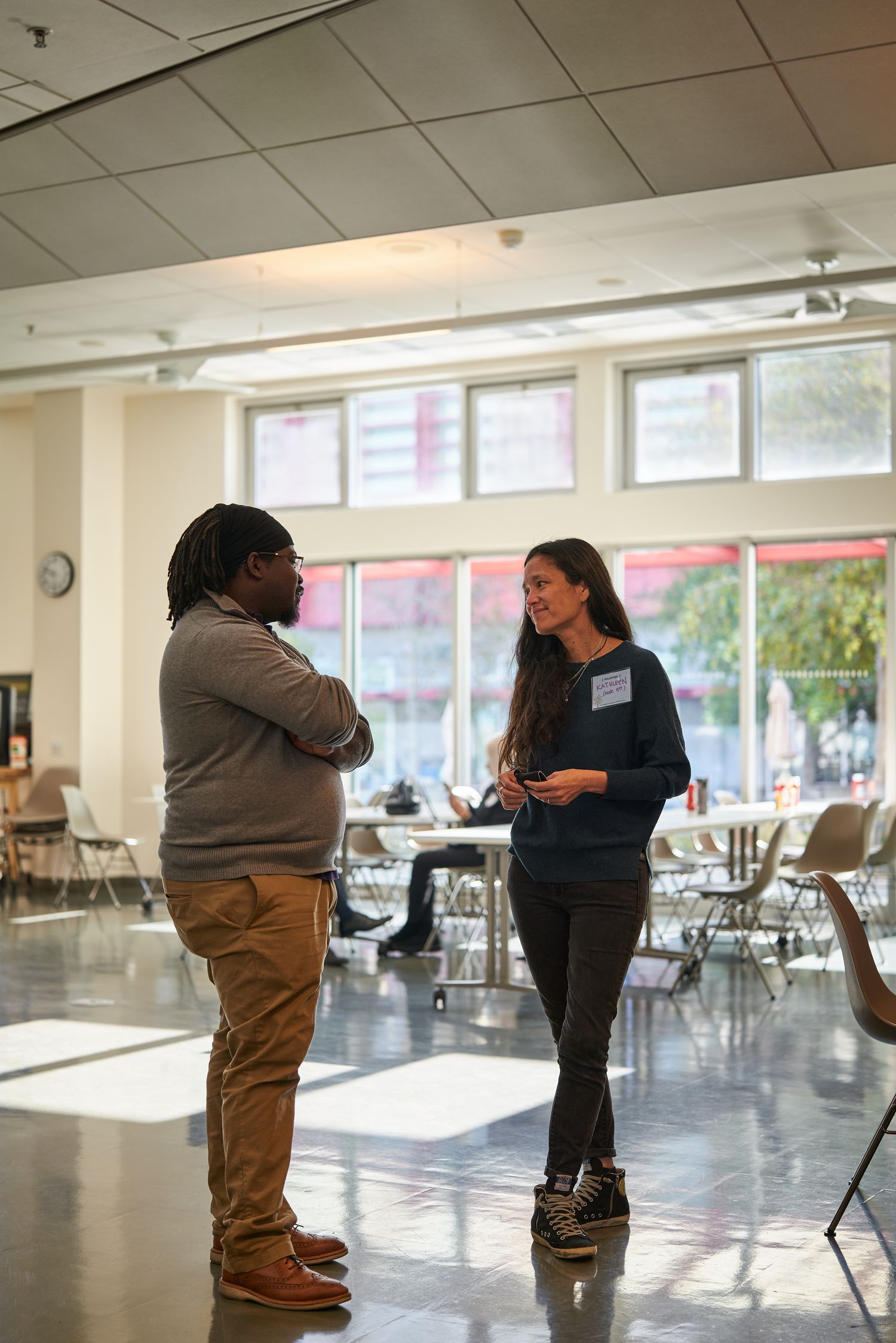 Two people having a conversation in a bright community center with chairs and windows.