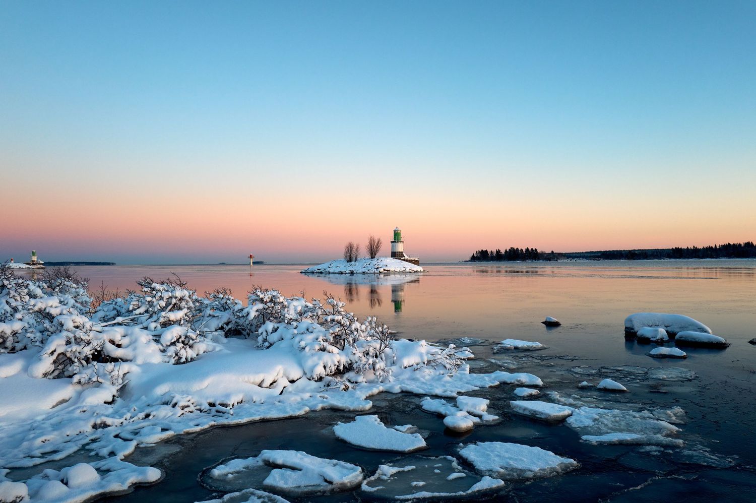 Frozen winter shoreline at sunset with ice formations and snow-covered trees silhouetted against pink sky.