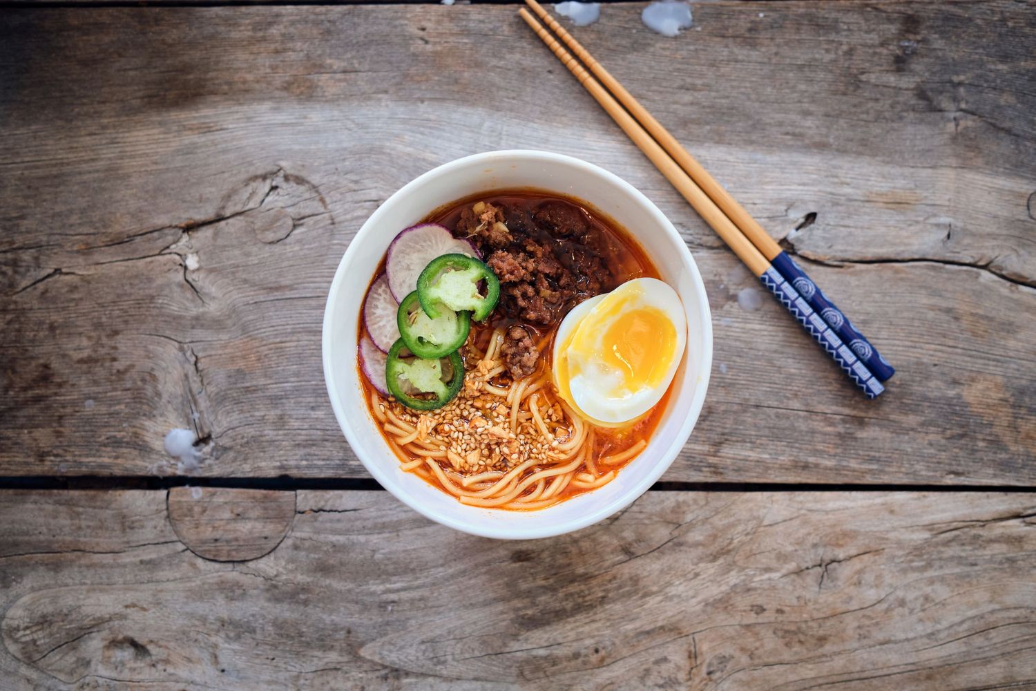 A bowl of ramen noodles with egg, cucumber and broth on a rustic wooden table.
