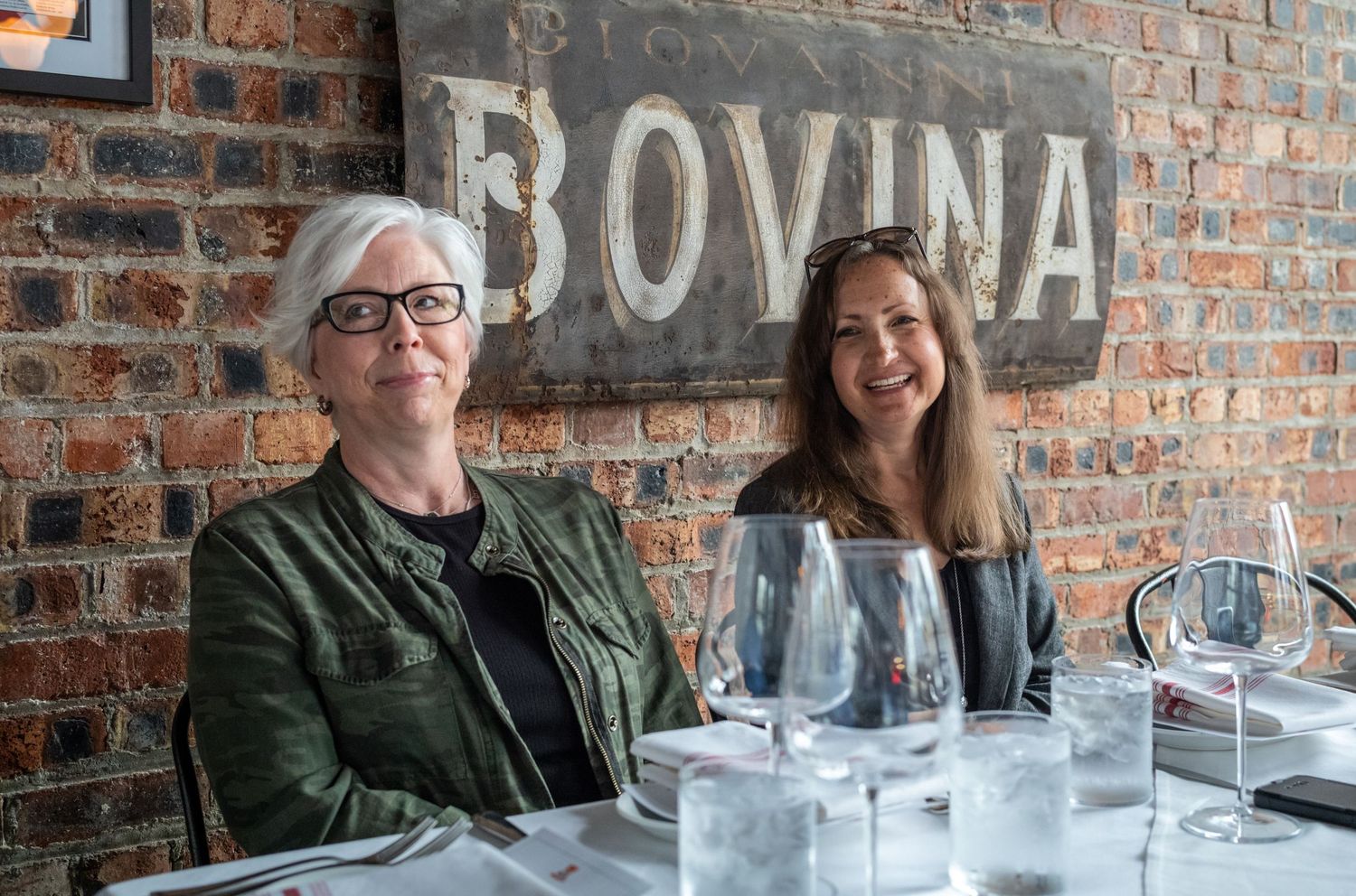 Two patrons seated at a restaurant table with wine glasses under a Bovina sign on exposed brick wall.