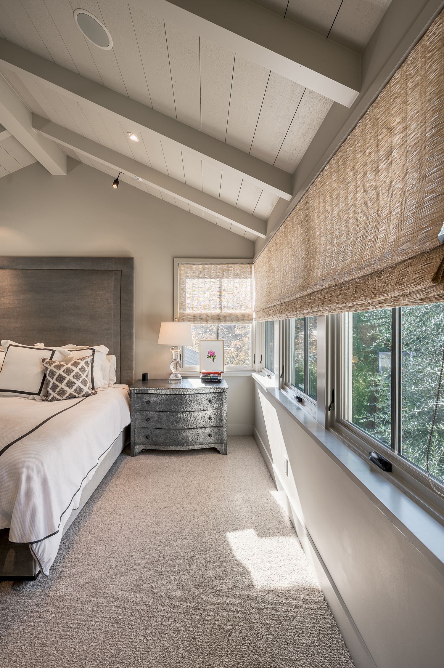 Serene bedroom with vaulted ceiling, woven window shades, and gray dresser beside a large window wall.