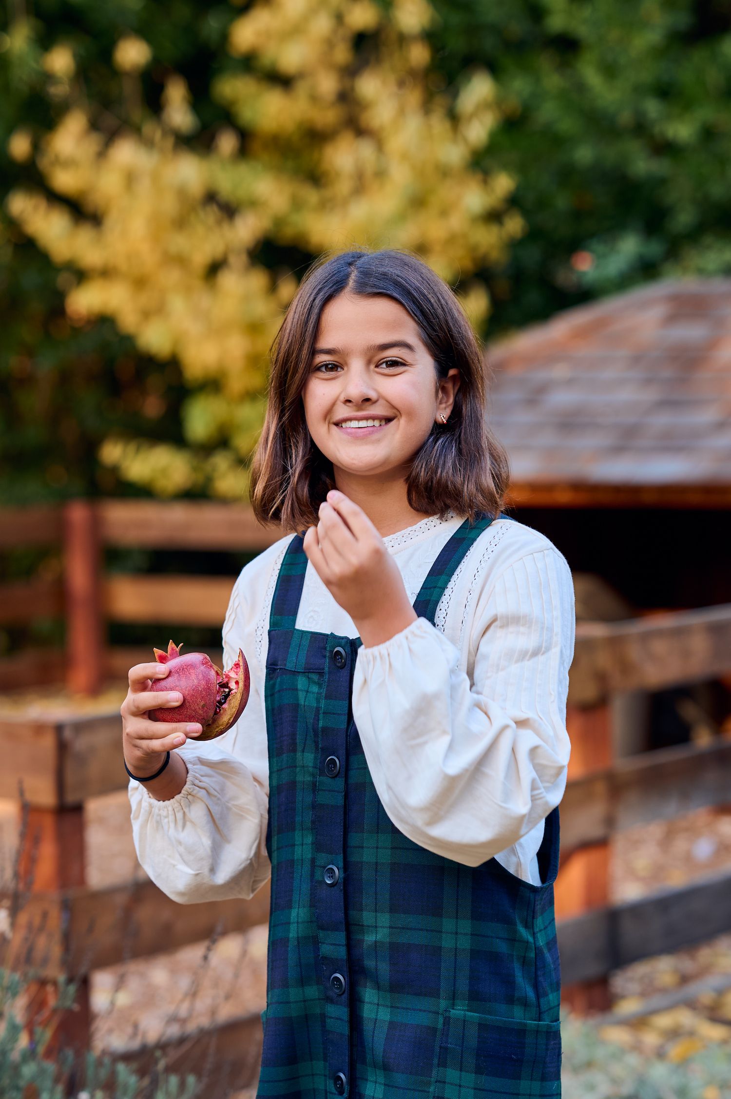 A person in a plaid jumper dress holds a red apple while standing in an autumn garden.