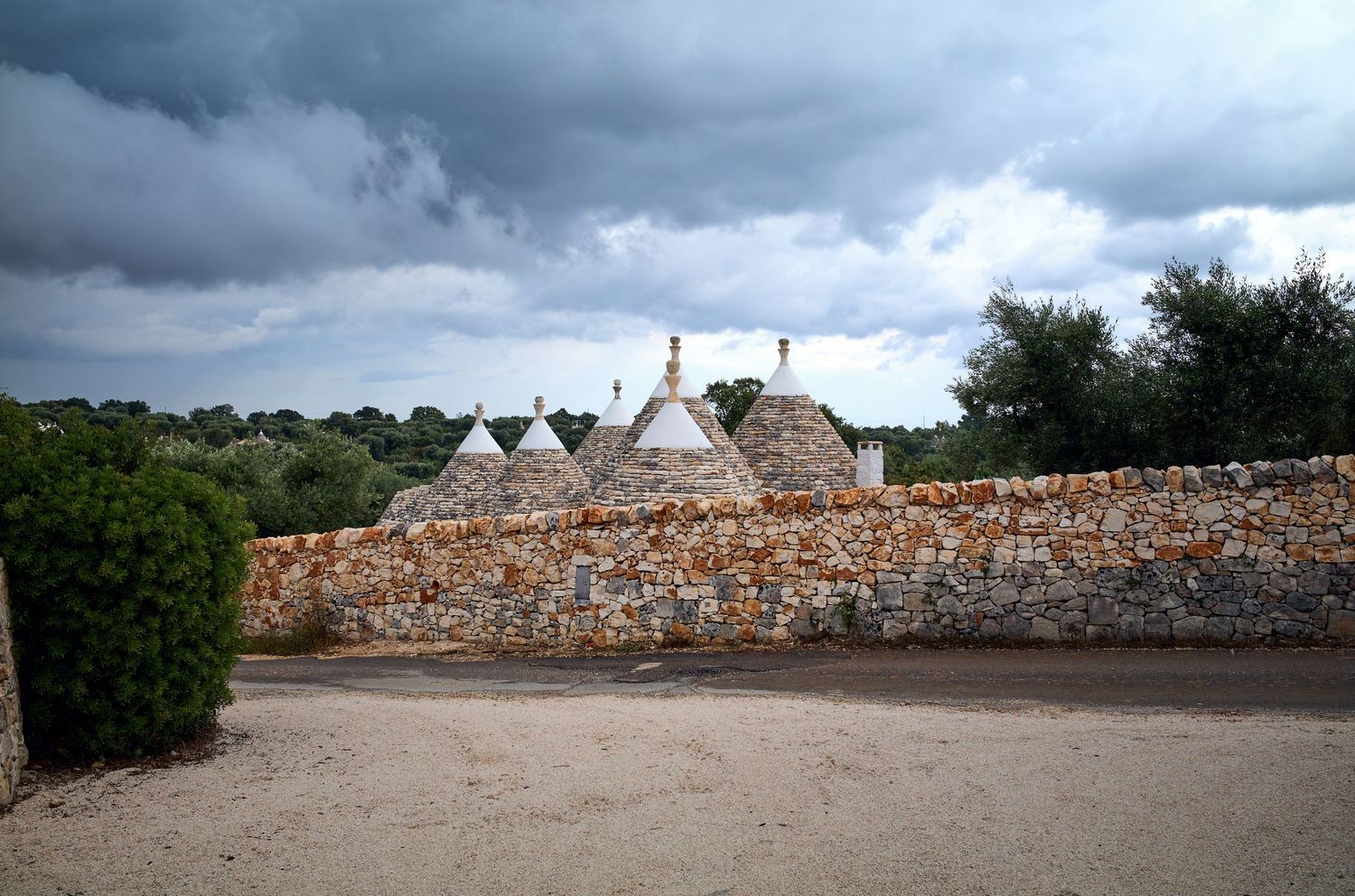 Traditional stone trulli houses with conical roofs behind a rustic wall under dramatic cloudy skies.