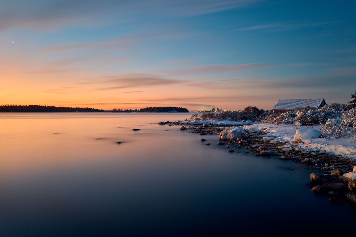 Snowy coastal landscape at sunset with calm waters reflecting golden sky and ice-covered rocks along the shore.