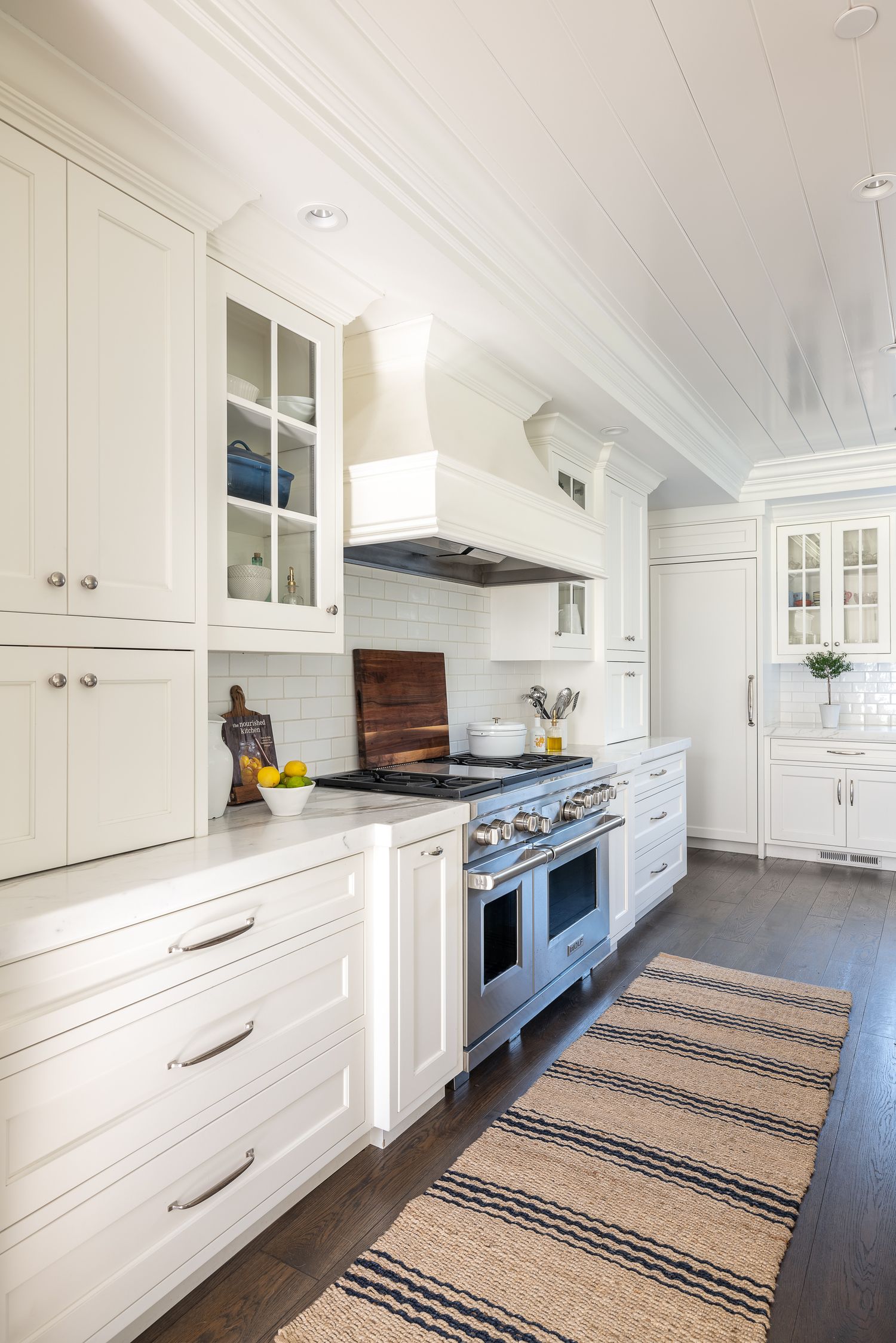 White kitchen with glass-front cabinets, professional range, and striped runner rug on dark hardwood floors.