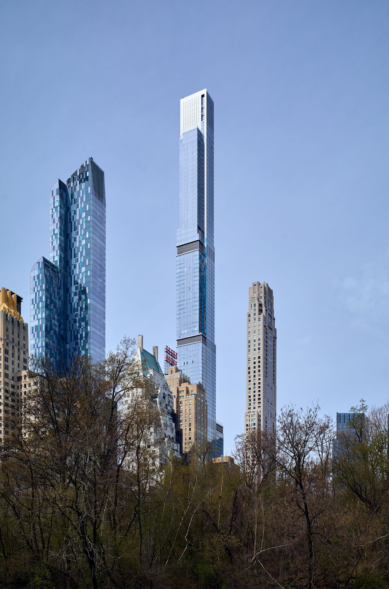Tall modern skyscrapers rise above Central Park trees against a clear blue sky in New York City.