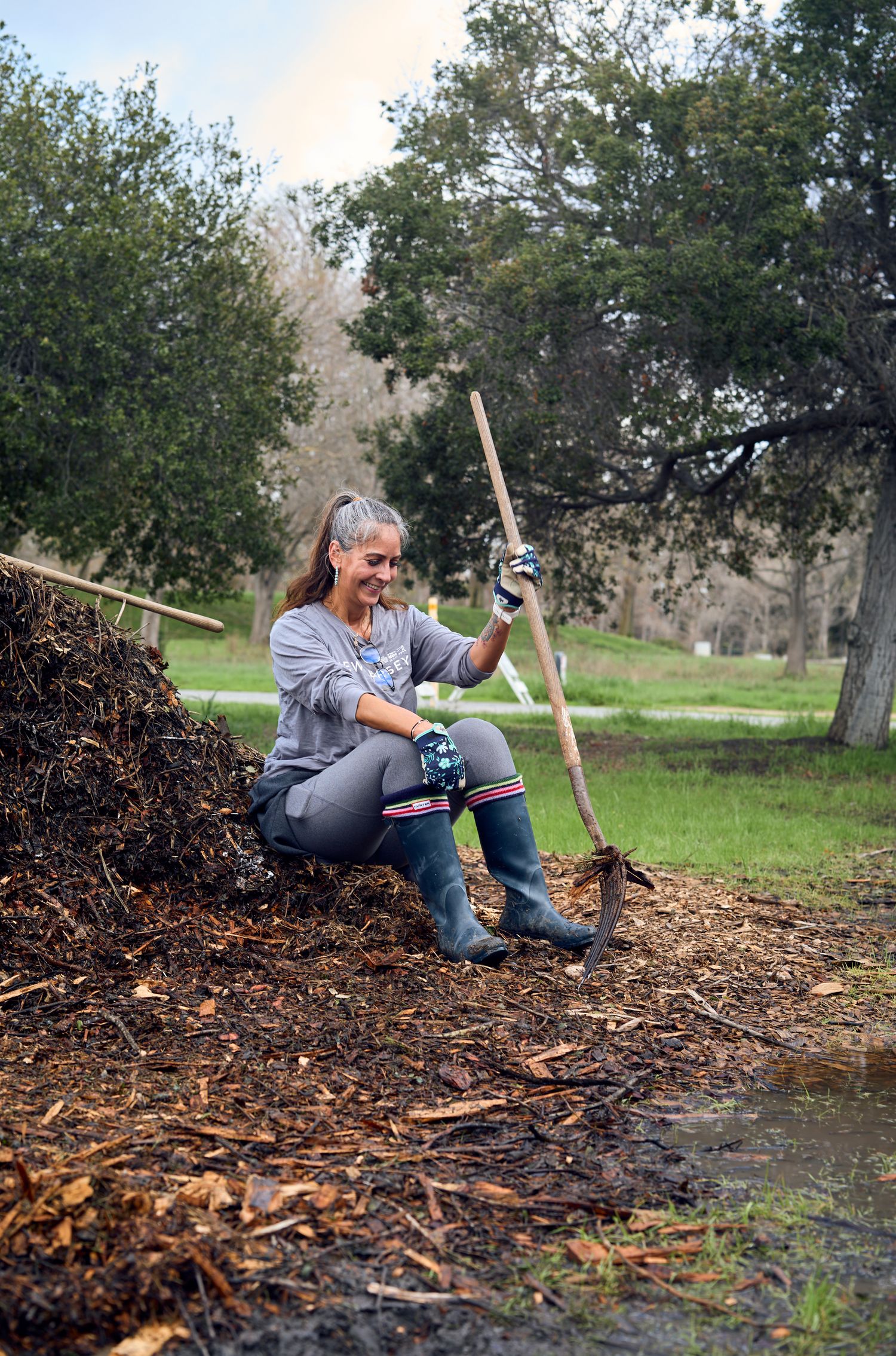 Gardener working with tools while sitting on mulch pile in park setting.