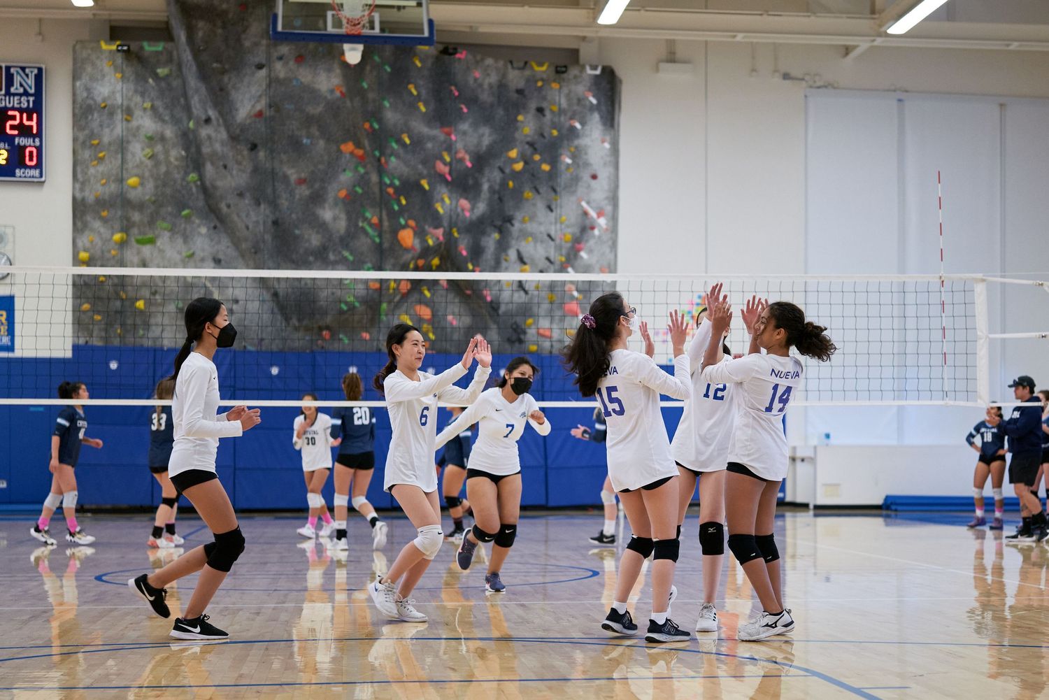 Female volleyball players celebrate together after scoring a point in a gymnasium with a climbing wall.