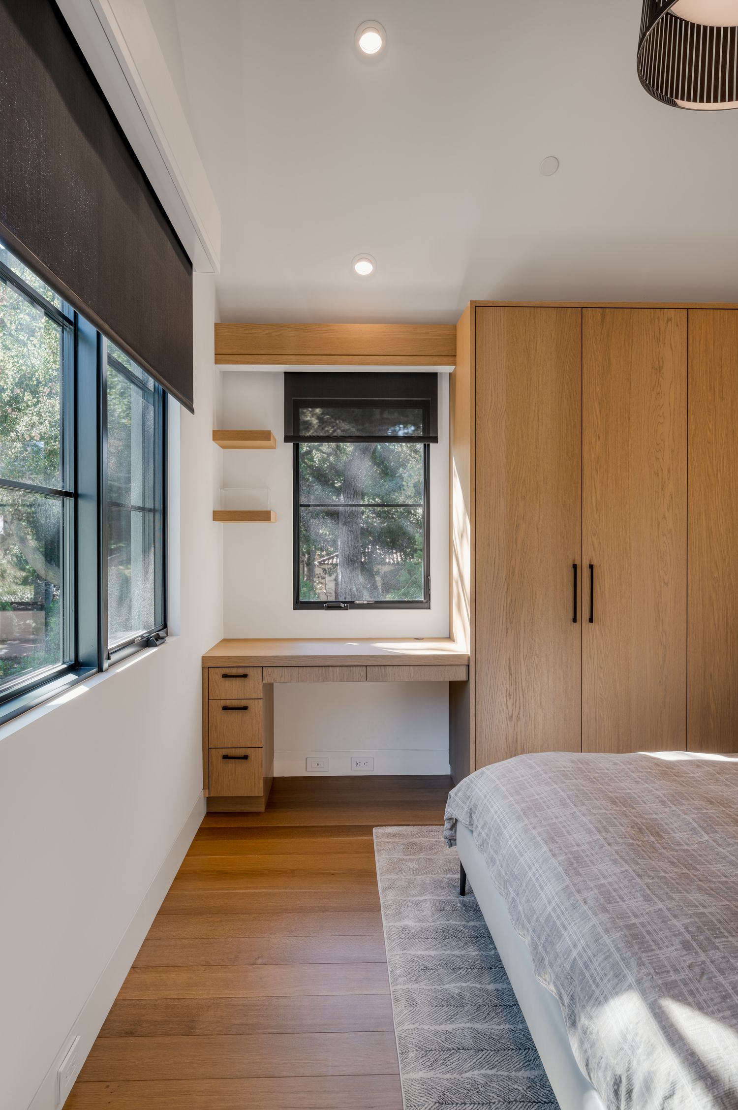 Modern bedroom features built-in wooden desk, windows, and hardwood floors in a minimalist design.