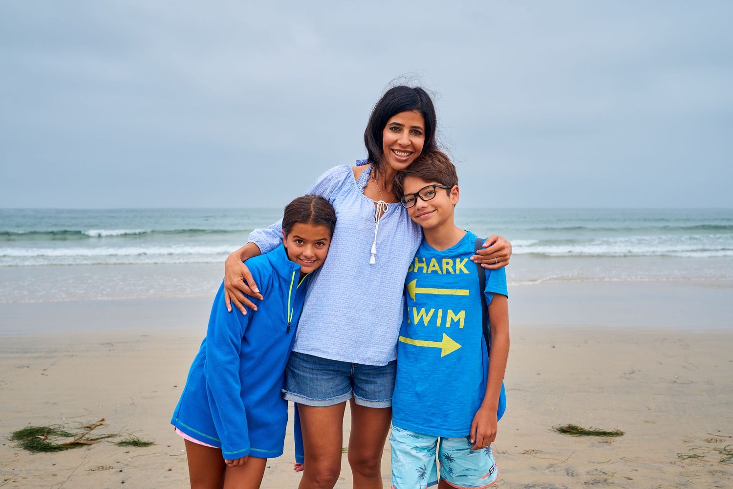 Three people stand together smiling on a cloudy beach with the ocean waves visible in the background.