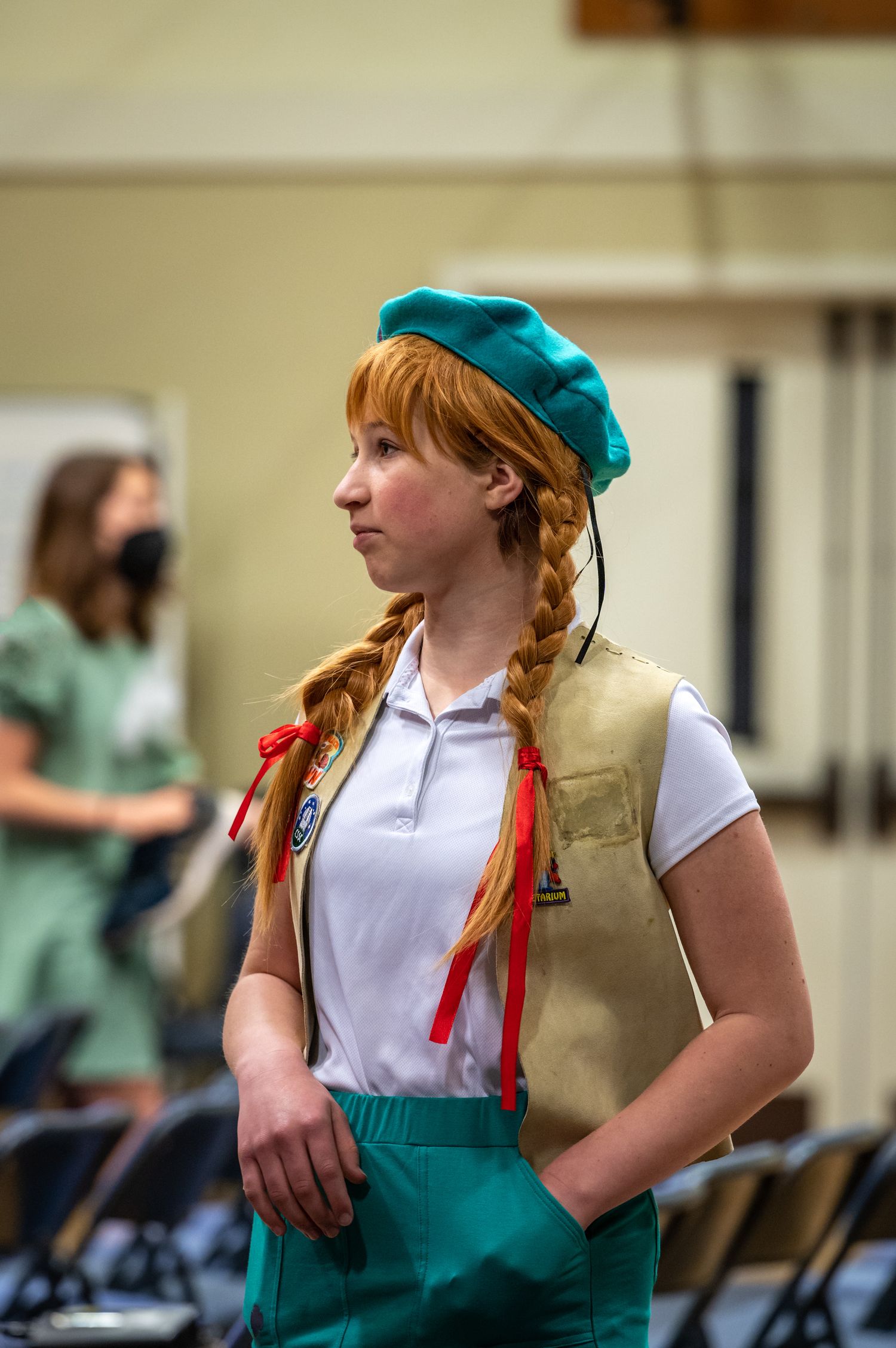 A person in a Girl Scout uniform with braided hair and teal beret stands in profile.