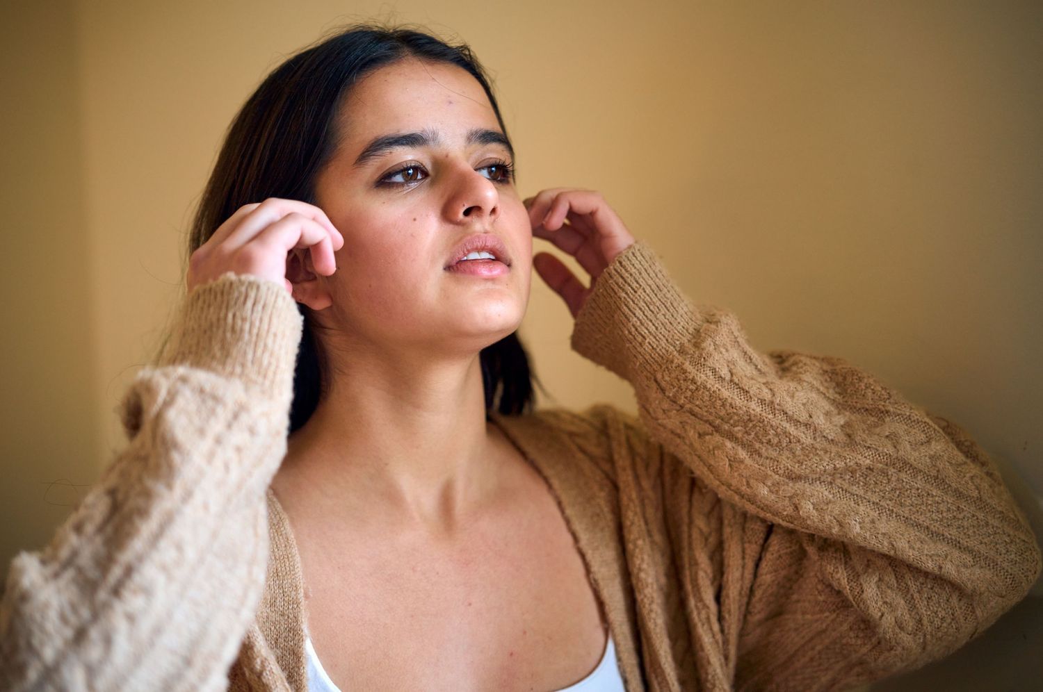 Person in beige sweater touching their hair against warm-toned background.