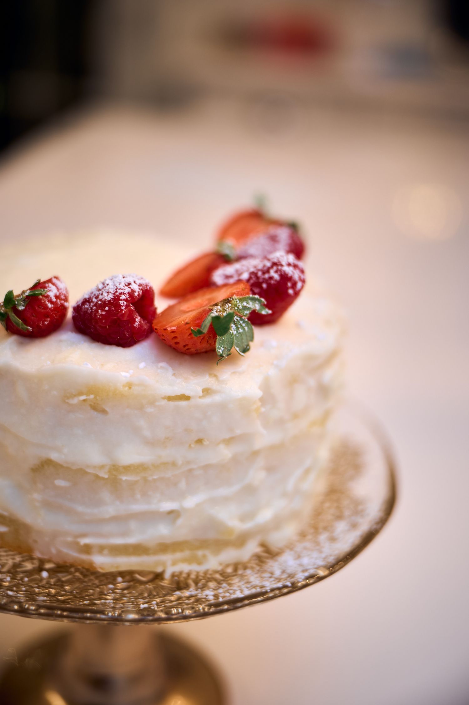 White layered cake topped with fresh strawberries on a gold cake stand, photographed in soft focus.