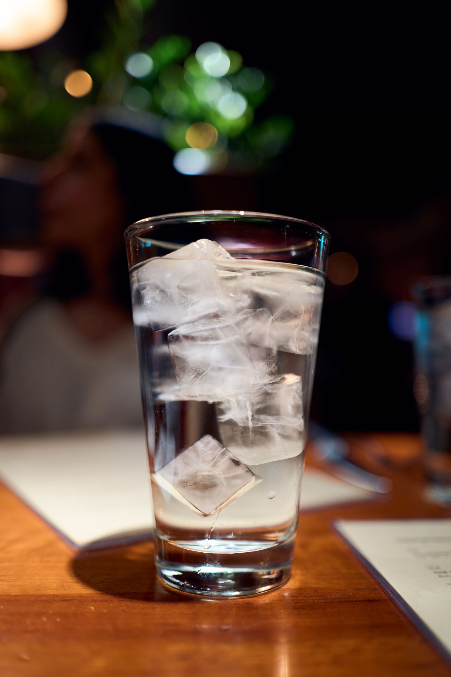 A tall glass filled with ice cubes on a wooden table with bokeh lights in the background.