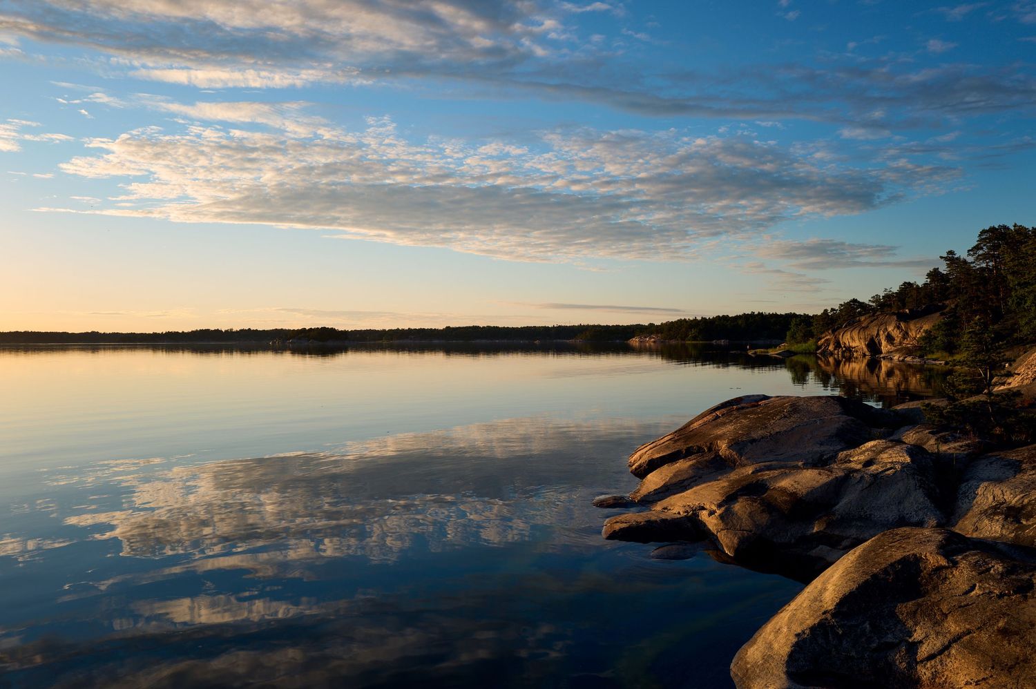 Calm lake reflecting sunset clouds with rocky shore and pine trees in foreground.