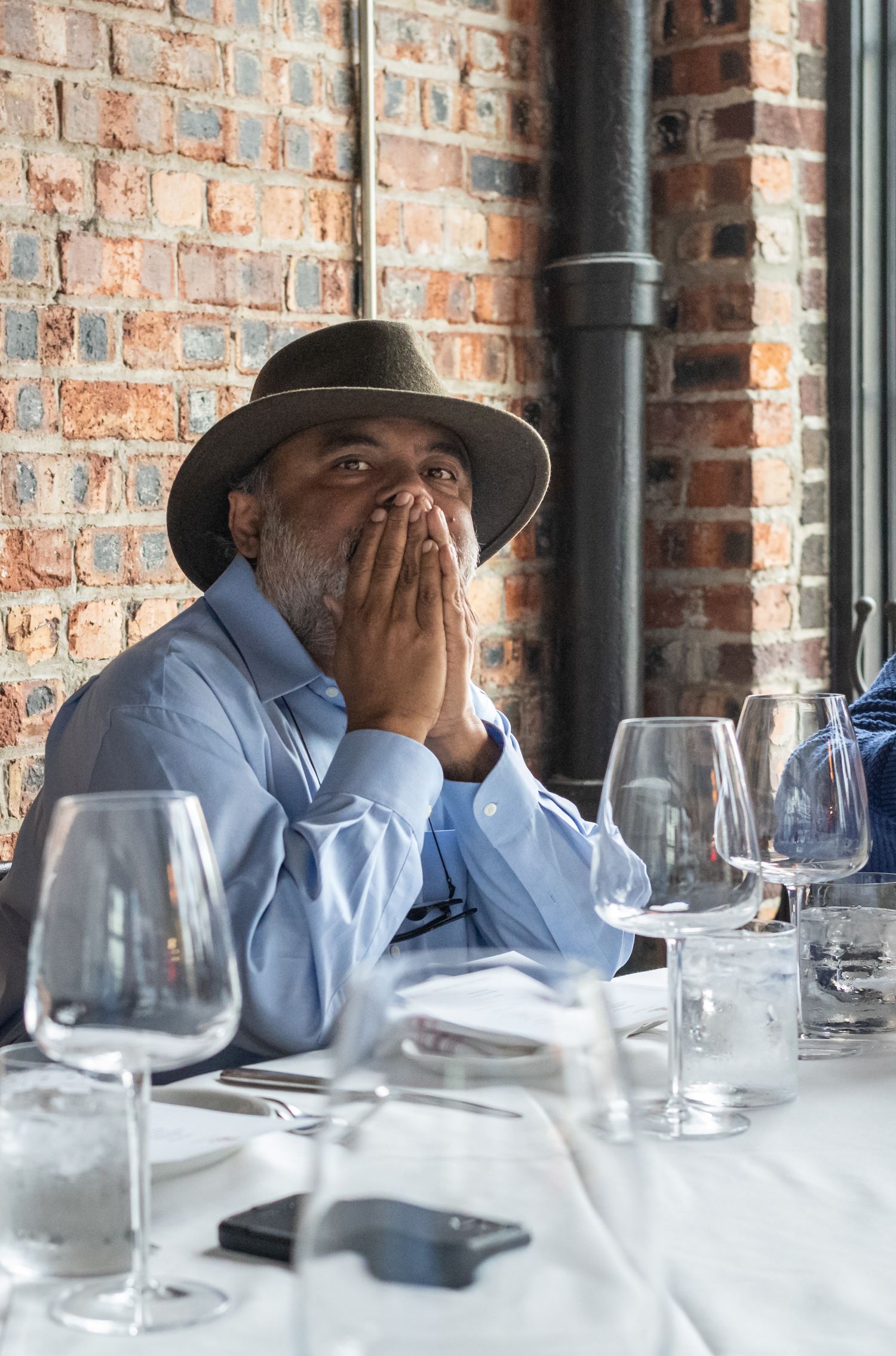 A person in a hat and blue shirt dining at a restaurant with wine glasses on the table.