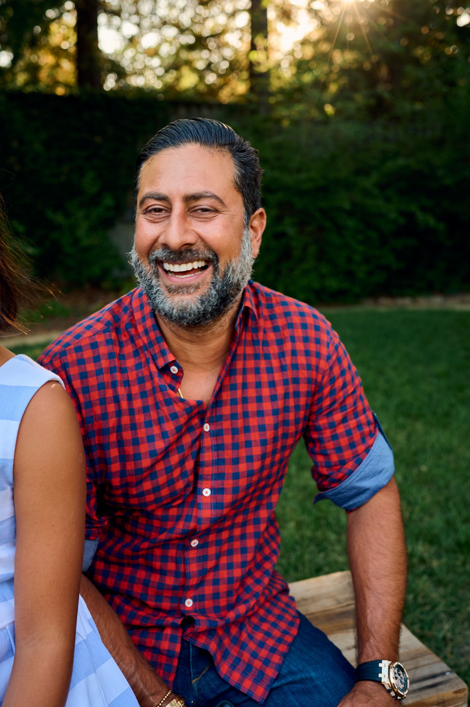 A person in a red and blue plaid shirt smiles while sitting outdoors at sunset.