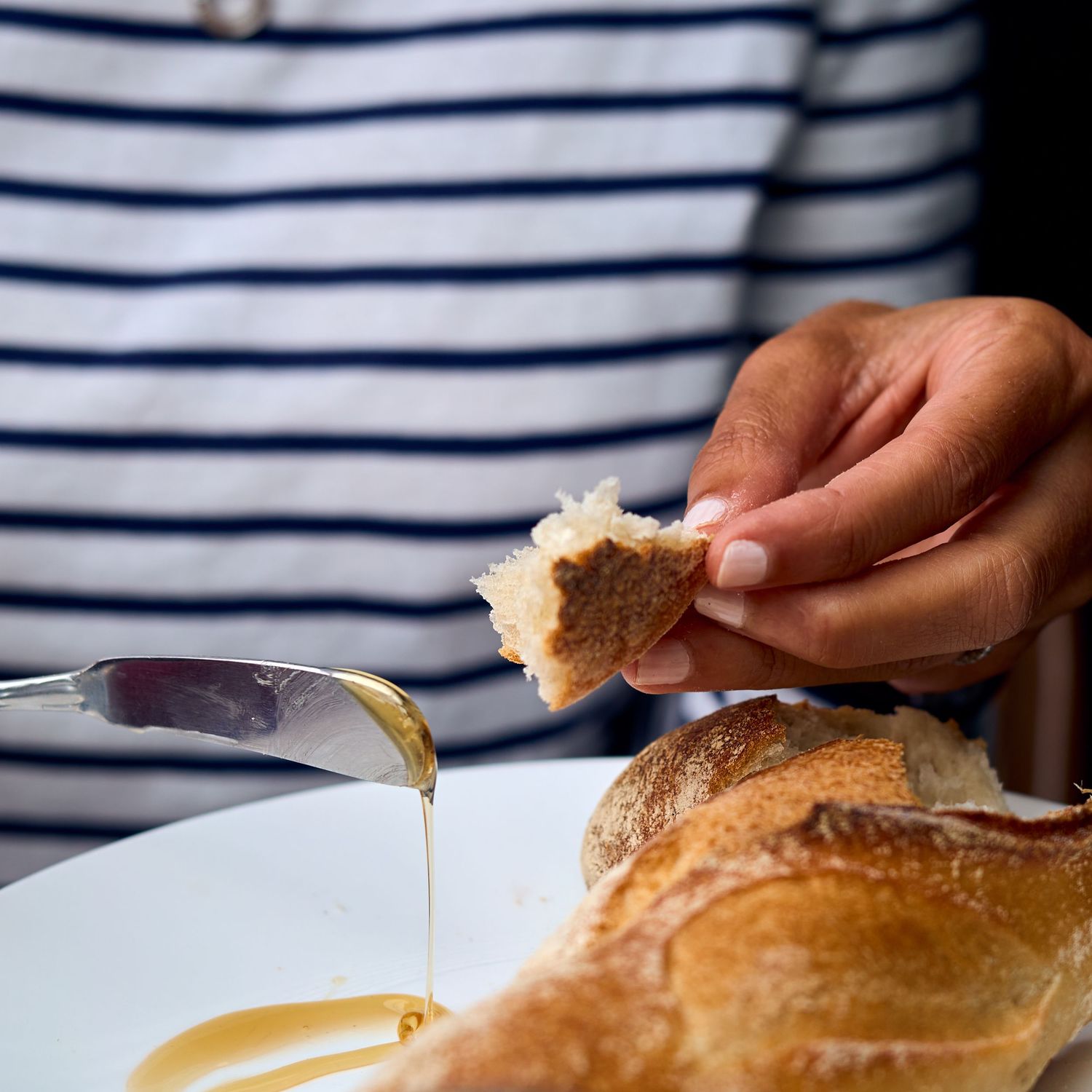 Hand drizzling honey over torn bread against striped fabric background.