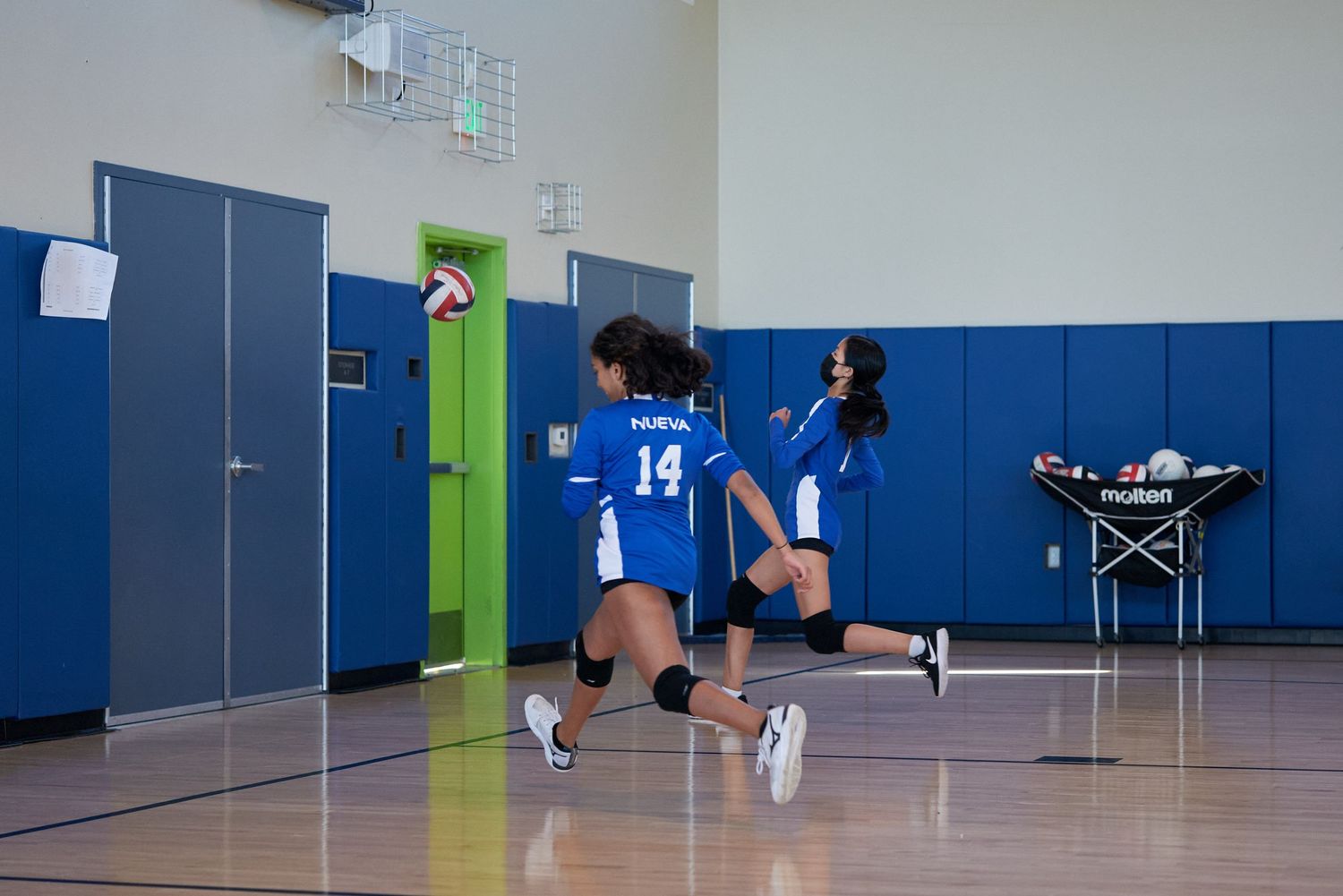 Volleyball players practicing serves in blue and white gymnasium.